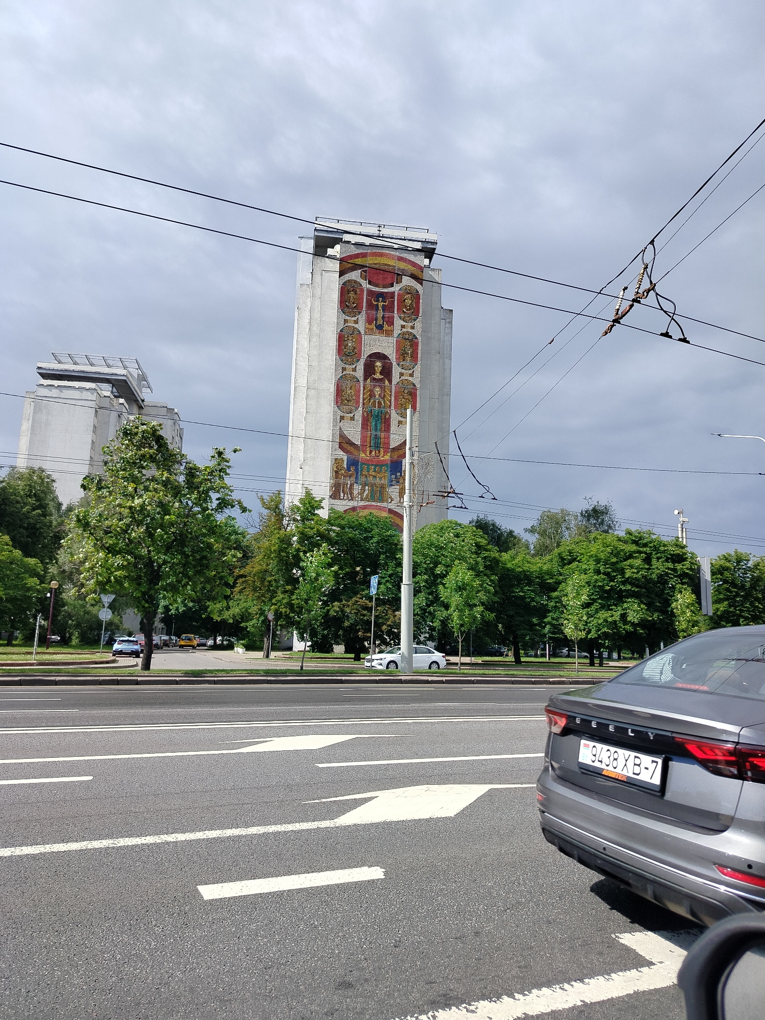 A street-level view shows a tall, narrow building with a large mosaic on its facade. The mosaic depicts a series of circular frames, each containing a figure. The building is light-colored and appears to be modern, with a flat roofline. Two similar, but smaller, buildings are visible to the left of the main building. The scene is viewed from a street, with a grey car in the foreground on the right. The car has a license plate that reads "9438 XB-7". The street is asphalt and has white lane markings. Trees line the street and a cloudy sky is visible above. There are overhead wires suspended across the scene.