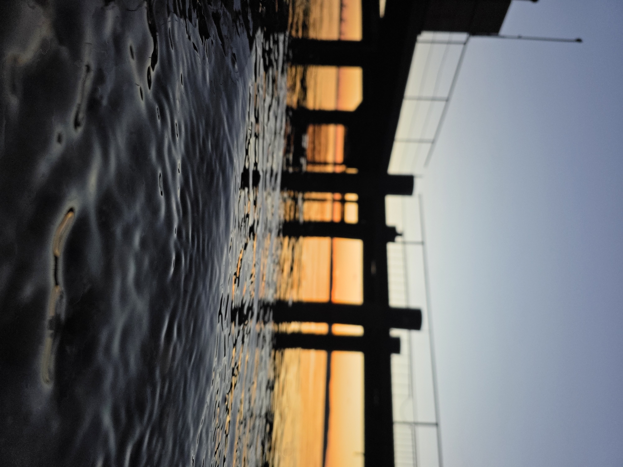 The image depicts an evening scene at the Kama River in Chaikovsky, Russia, as observed from the water level. The foreground is dominated by dark, rippling water reflecting the colors of the setting sun. A wooden pier or dock stretches horizontally across the image. Its dark silhouette is punctuated by gaps allowing views of the orange and yellow light from the sunset reflecting on the water's surface. The sky transitions from shades of orange near the horizon to pale blue above the pier.