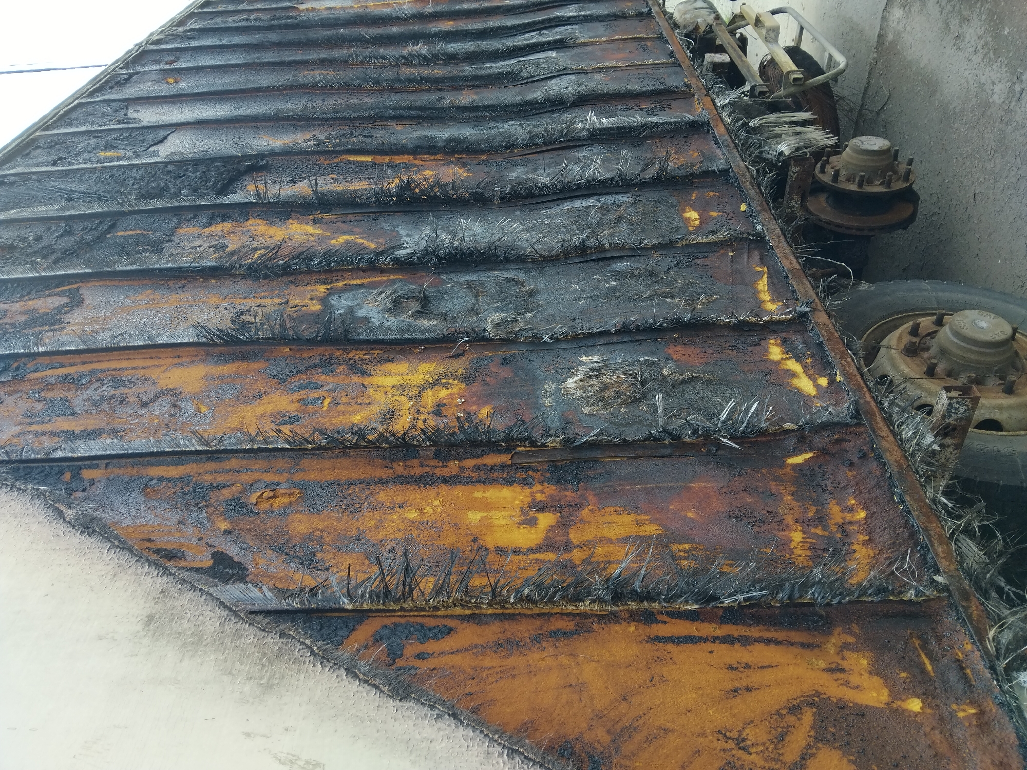 The image shows the charred side of a truck trailer. The trailer's exterior is largely covered in black soot and melted material, with patches of orange and yellow paint visible beneath. The surface is uneven and textured, with some areas appearing melted or bubbled. The vertical corrugated panels of the trailer are clearly visible, warped and discolored by heat. The underside of the trailer, including wheels and suspension components, is also visible and appears to be dirty and somewhat damaged. The ground beneath the trailer is a concrete or asphalt surface, showing some debris and discoloration. The scene is devoid of any people or immediate signs of ongoing activity, suggesting a post-incident state. The image is a close-up, focusing on the damage to the trailer. There is no contextual information within the image that specifies the location as Kyiv, Ukraine.