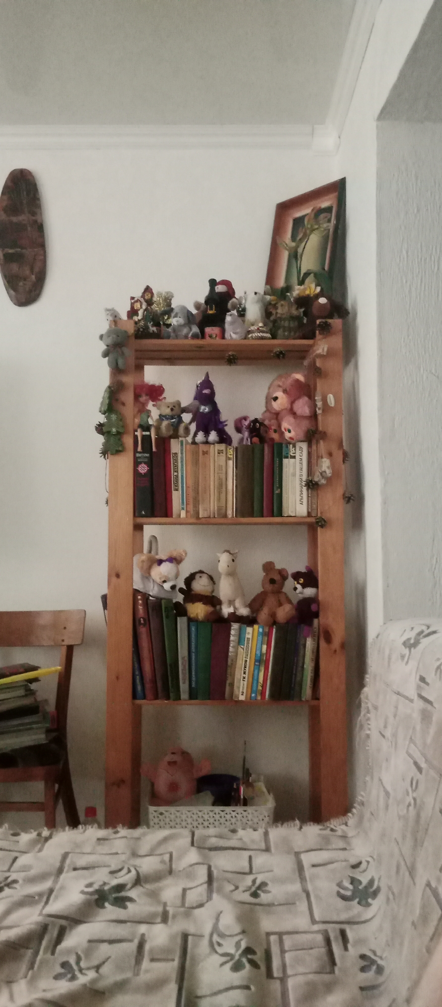 The image shows a wooden bookshelf filled with various stuffed animals and books. The bookshelf is tall and has multiple shelves. On the top shelf, there are several small stuffed animals, including what appears to be a teddy bear, a gray dog, and some other figures. The second shelf from the top also holds stuffed animals, including a pink teddy bear, a purple creature, and a smaller stuffed dog. The third shelf has more stuffed animals, such as a white dog with a bow, a small brown monkey-like toy, a white horse, and a brown teddy bear. The bottom shelf contains a pink stuffed toy with its arms up, sitting in front of a white basket filled with various items.
The books on the shelves are mostly lined up vertically, with their spines facing outwards. Some of the book spines have visible text, but it is mostly illegible due to the image quality and resolution. The colors of the book spines vary, including red, green, brown, and white.
To the left of the bookshelf, there is a wooden chair with a stack of books on it. In the background, the wall is white, and there is a framed picture hanging on the wall above the bookshelf. The overall scene appears to be indoors, likely in a living room or a den, given the presence of the bookshelf, chair, and stuffed animals, which suggest a home environment. The lighting suggests it is daytime, but no specific time can be determined. The overall mood is casual and cozy, with a focus on collection and display of toys and books.