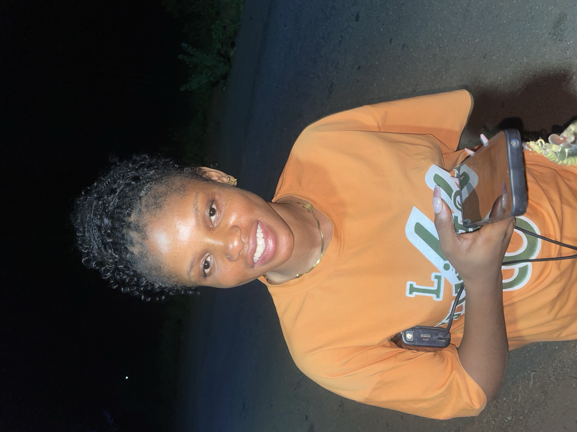 A woman with curly hair is outdoors, holding a smartphone in her right hand and a portable charging device in her left. She is smiling. She is wearing a gold chain and a bright orange T-shirt with a large green and white design. The location appears to be an outdoor paved area with vegetation visible in the background. The lighting is dim. Based on the prompt, the location is Ogbe, Nigeria.