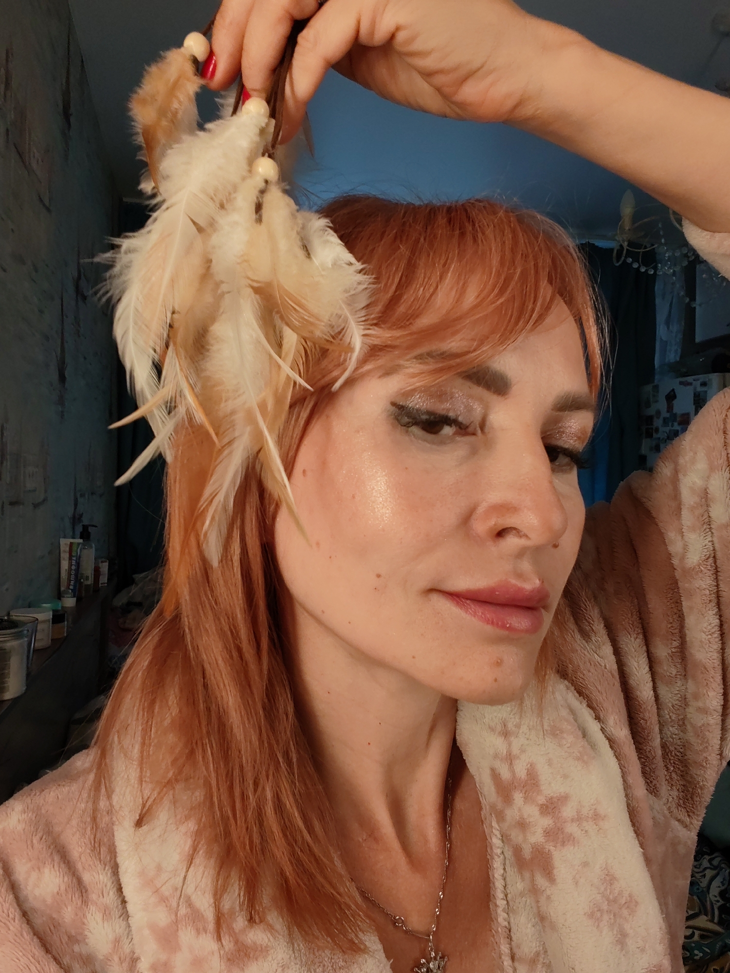 A woman with reddish-brown hair, wearing a fluffy pink bathrobe, is holding a feather accessory above her head. She has on makeup, including eyeshadow and eyeliner. The background appears to be an indoor setting with a blue-toned wall and some shelves with bottles. The image is a close-up shot focusing on the woman's face and the feather accessory.