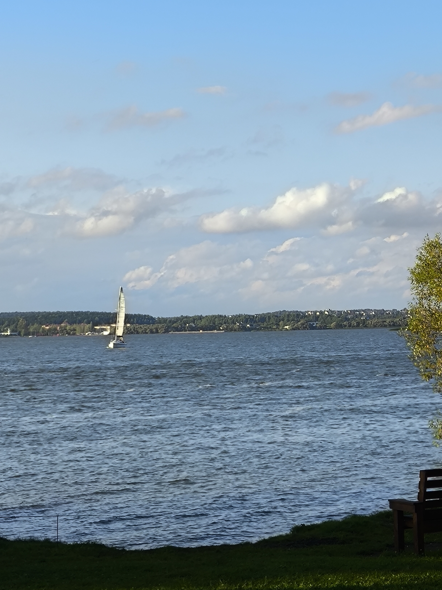 A wide shot captures a tranquil lake under a partially cloudy sky. In the distance, a lush green shoreline is dotted with buildings, suggesting a settlement within the Ждановіцкі сельскі Савет, Belarus. A lone sailboat glides across the rippling water, its white sail catching the sunlight. In the foreground, a dark wooden bench sits on a grassy bank, offering a vantage point to observe the serene landscape. The scene is bathed in the soft light of what appears to be late afternoon or early evening.