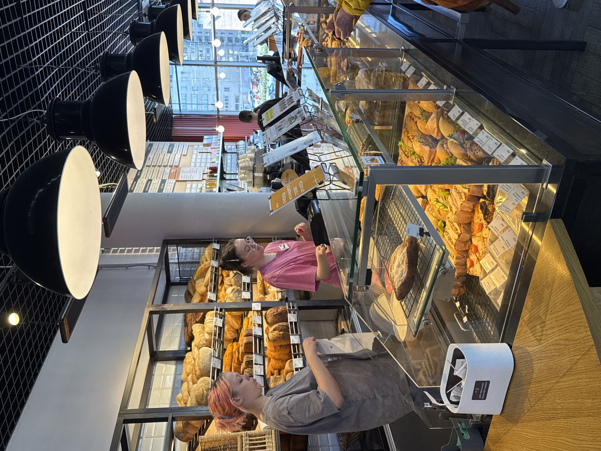 This image depicts the interior of a bakery. Two women are near the display cases filled with bread and pastries. One woman has pink hair and is wearing a gray shirt, while the other is wearing a pink shirt. A glass display case contains various sandwiches and pastries. Several black dome lights hang from the ceiling. In the background, a window shows a cityscape. According to the information included within the image metadata, it was taken in Saint Petersburg, Russia.