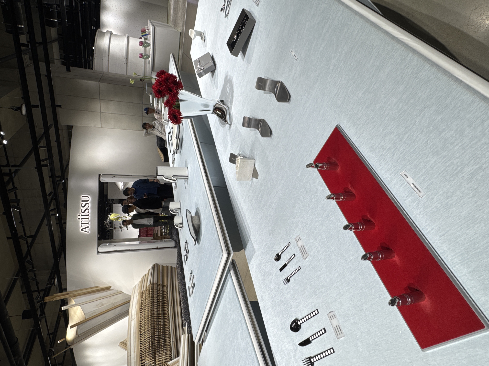 The image captures an interior exhibit at the Atiissu store, potentially located in Seoul, South Korea, as indicated by the store’s name displayed. The exhibit features a long, pale blue table showcasing a variety of metal objects and utensils, some arranged on a rectangular red panel. The objects include what appear to be decorative elements and possibly functional items like cutlery. There is also a white vase filled with red flowers positioned towards the center of the table.
In the background, several people are seen browsing the store. The environment suggests a well-lit, modern interior space, with structural architectural elements and installations contributing to the overall design of the store.