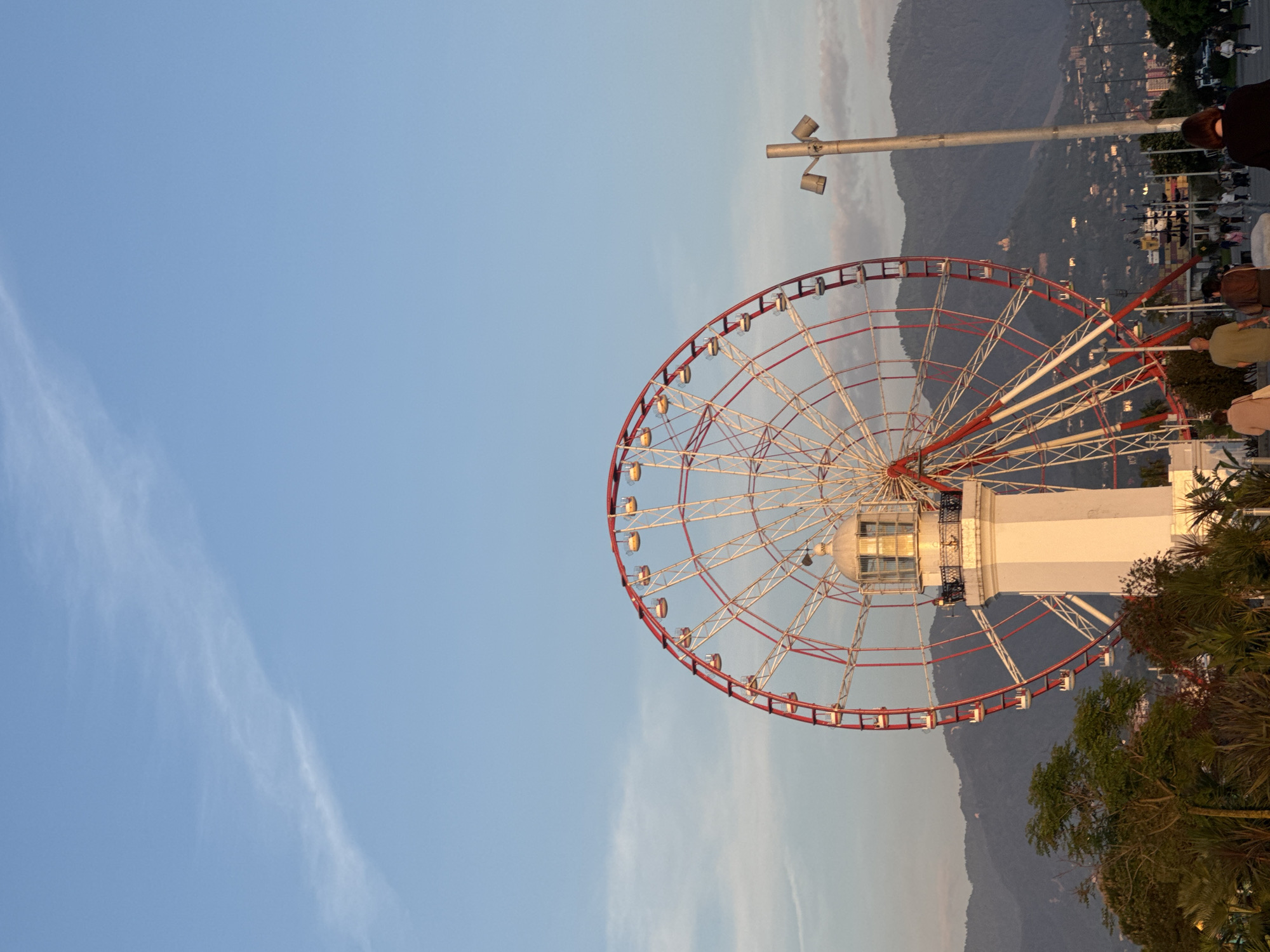 Here is a description of the image:
The image shows a Ferris wheel with a red and white frame. The wheel appears to be mounted on a white structure. The sky is visible in the background, with a clear blue color and some wispy clouds on the left. In the upper right corner, there are some blurred figures of people and a light post, with a backdrop of mountains and city lights. A small portion of greenery is visible at the bottom right of the image. The image appears to be rotated, with the Ferris wheel angled.