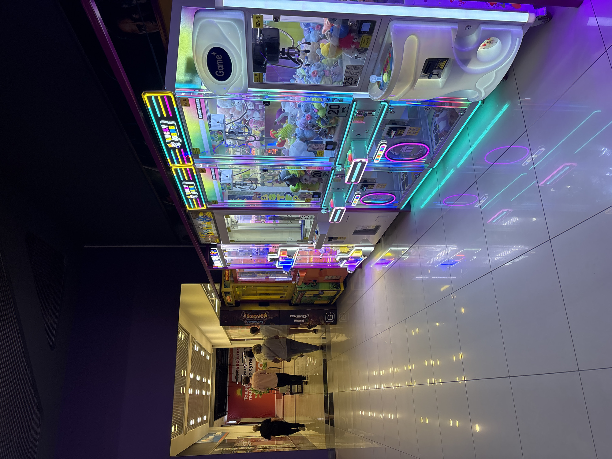 The image depicts an indoor shopping mall interior in Naberezhnye Chelny, Russia, characterized by a brightly lit hallway with tiled floors. The scene focuses on a row of illuminated arcade games, specifically claw machines, displaying an assortment of plush toys. In the background, several individuals are walking down the hallway. The arcade machines are adorned with neon lights in shades of blue, pink, and yellow, creating a vibrant and colorful atmosphere. The flooring is highly reflective, mirroring the lights of the arcade machines and the overhead lighting. The overall environment suggests a commercial entertainment space.
