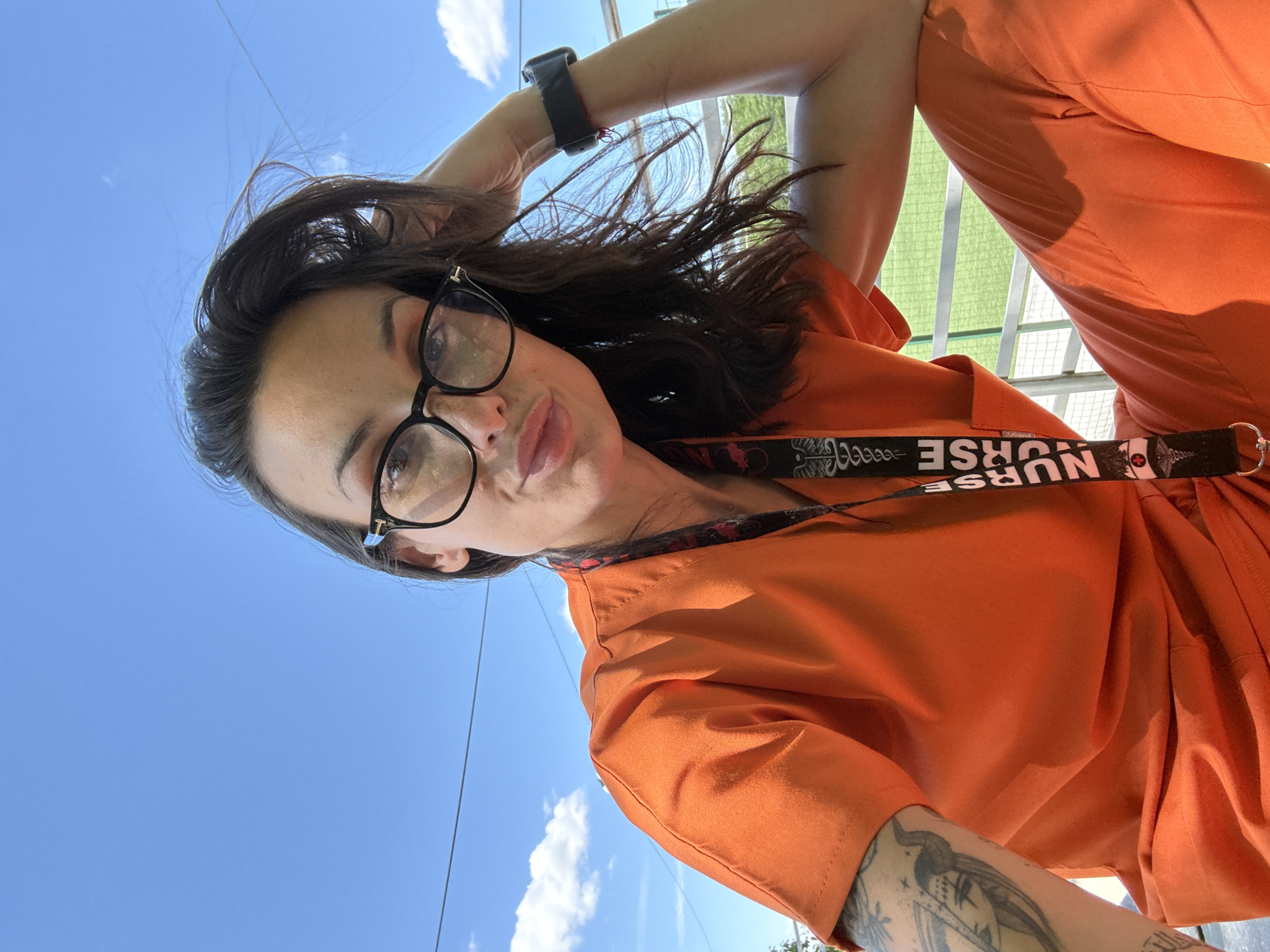 A woman with long, dark hair and glasses, wearing bright orange scrubs and a lanyard that reads "NURSE," is posing for a selfie outdoors on a sunny day. She has tattoos on her left arm and is wearing a watch on her right wrist. The background shows a clear blue sky with some clouds, a white railing, and a green field, possibly a sports field, suggesting a location like a hospital, clinic, or outdoor recreational area. The image does not provide specific details to identify the location in Pleven, Bulgaria.
