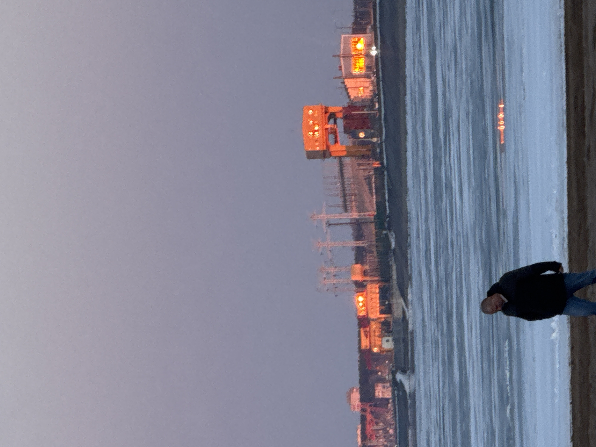 A long shot depicts a body of water, likely a river, adjacent to an industrial area in Noviy, Russia. The sky is overcast. In the foreground, a single male stands near the water's edge. He is wearing a dark jacket and jeans. In the background, industrial cranes and facilities are visible, lit up against the dusk sky. High-voltage electrical towers run from the horizon down the shore. The ground is covered in sand and ice.