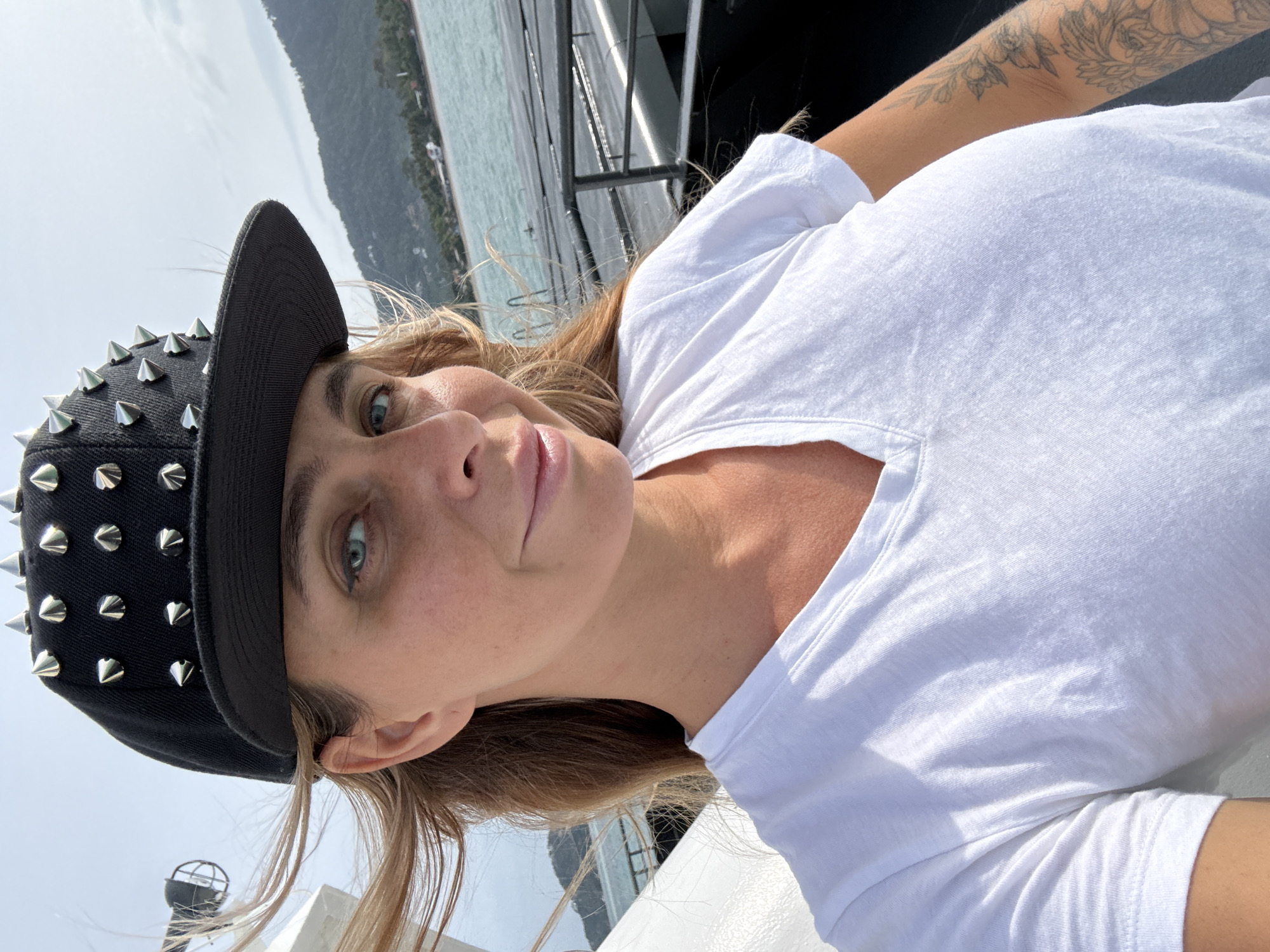 A woman in a white V-neck t-shirt and a black baseball cap with silver studs is taking a selfie on a ferry. Her blonde hair is blowing in the wind. The background shows a body of water, a distant mountainous island with buildings, and the deck of a ferry. The location is on a ferry in the Ko Samui District, Thailand.