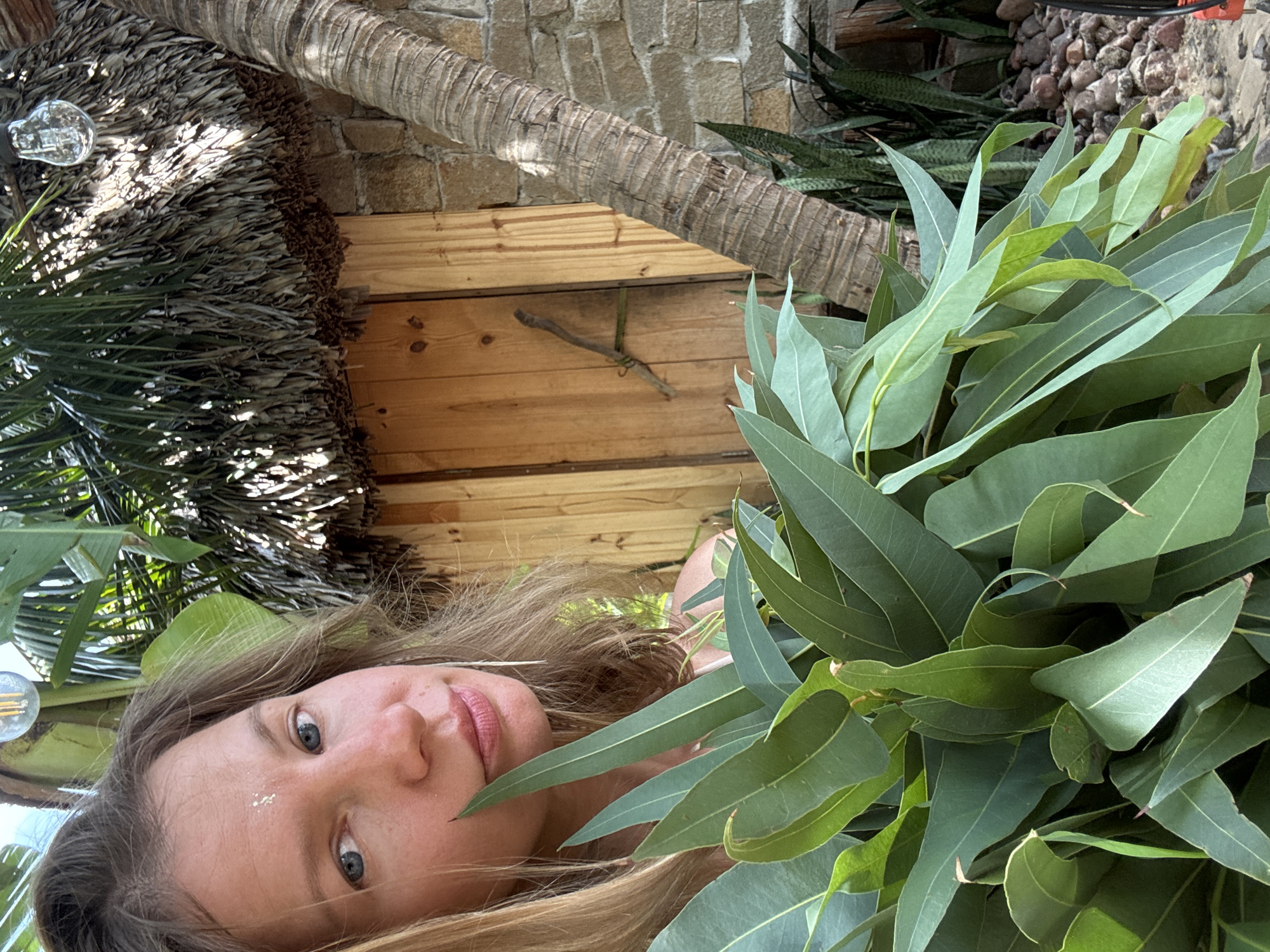 A medium-close-up, eye-level, outdoor selfie shows a woman with blonde hair and blue eyes holding a large bunch of green eucalyptus leaves. She is looking directly at the camera with a slight smile and has a small silver decoration on her forehead. In the background, there is a thatched roof structure, a palm tree trunk, a stone wall, and a wooden door. The overall setting appears to be a tropical environment, possibly in Mũi Né, Vietnam, given the style of the thatched roof and surrounding vegetation. The lighting suggests it is daytime, with sunlight filtering through the leaves and casting shadows.