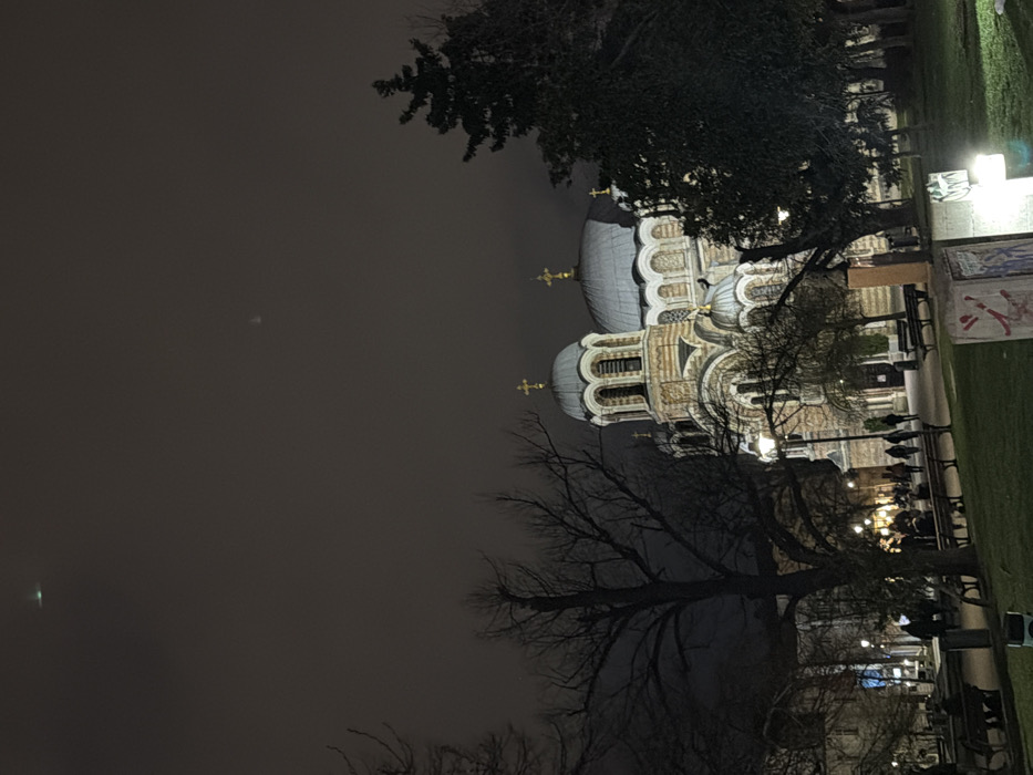The image displays the Sveti Sedmochislenitsi Church (Church of the Seven Saints) in Sofia, Bulgaria, at night. The illuminated church, a large structure with multiple grayish-blue domes topped with golden Orthodox crosses, features a distinct striped stone facade. In the foreground, a public park occupies the lower and mid-ground, characterized by green ground cover, bare deciduous trees, and several evergreen trees. Numerous park benches are present throughout the area, some occupied by indistinct human figures. Paved pathways are visible, along which additional human figures are walking. Street lamps provide artificial illumination, casting light on the park and surrounding areas. A concrete structure with visible graffiti is situated in the lower right foreground. The dark, cloudy sky contains two faint, distant light sources: a small green light in the upper left quadrant and a diffuse whitish glow in the upper right quadrant.