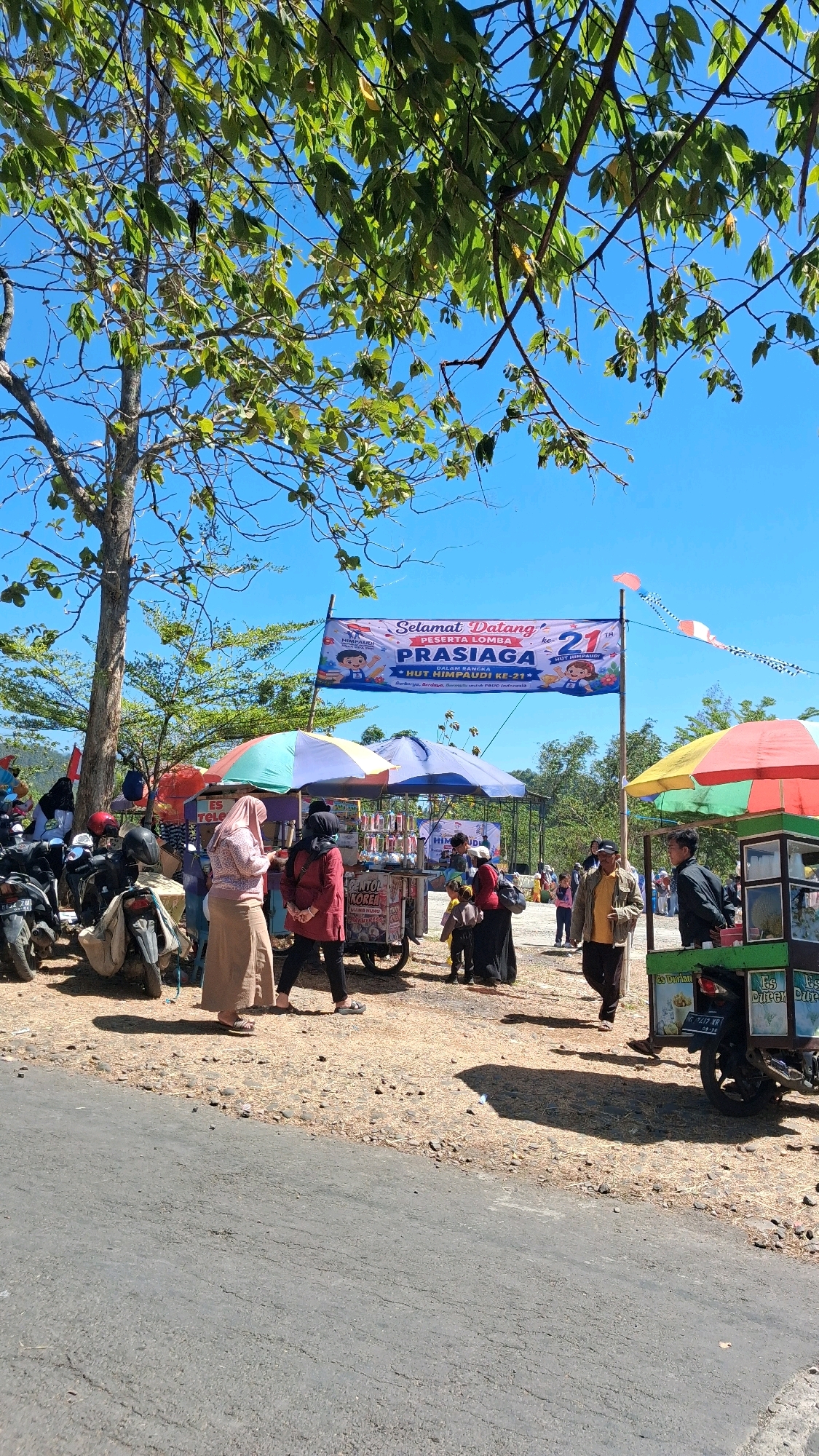 A group of people are gathered outdoors under a blue sky with some tree branches in the foreground. In the center of the image, a banner reads "SELAMAT DATANG PESERTA LOMBA PRASIAGA KE-21 HUT HIMPAUDI KE-21". To the left of the banner, several motorcycles are parked. In front of the banner, there are food stalls with colorful umbrellas. People are standing around the stalls, some appear to be customers and vendors. The ground is covered in dry grass and small stones. In the background, there are trees and a slightly hilly landscape. The overall scene suggests an outdoor event or festival taking place in a somewhat rural or park-like setting.