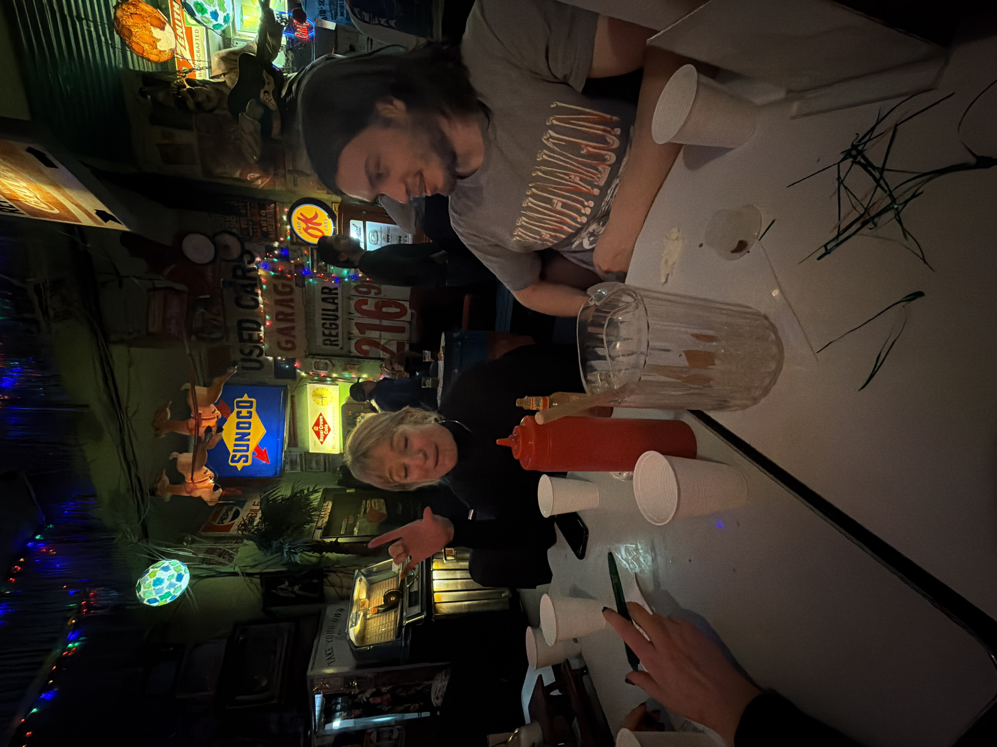 The image shows a group of people seated at a table in a dimly lit establishment. The location appears to be decorated with vintage signs and various lighting fixtures, suggesting a retro or themed setting, with the signs reading "USED CARS GARAGE, REGULAR 2169 AUTO" and "SUNOCO." The image was most likely not taken in the city of New York, United States of America, since the vintage gas prices are not common, and New York City does not have vintage garages with the signs visible in the image.
Three individuals are partially visible in the image. One person, a man with a beard, is seated at the table, wearing a grey t-shirt and a black headband. Another person is seated closer to the center of the image, a woman with blonde hair, who is seen gesturing with her hand. A third person is present on the right side of the image, with only their hands visible, clad in a black sleeve.
Several objects are present on the table, including cups, bottles, and other paraphernalia.