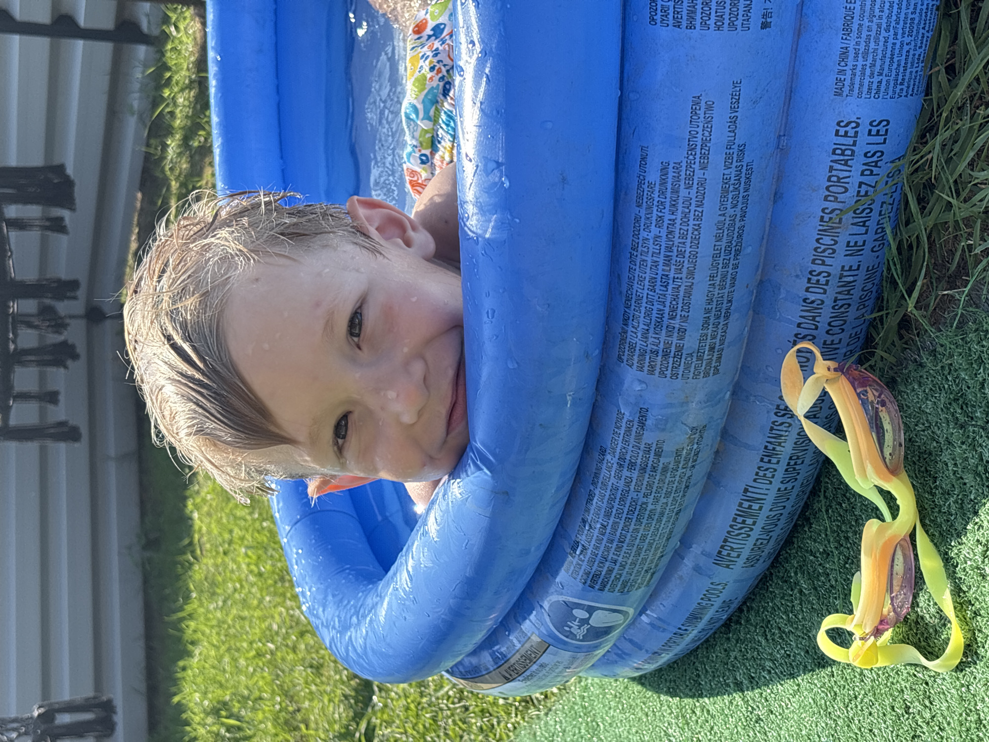 A young boy with wet blond hair smiles from an inflatable blue pool filled with water. He is wearing a colorful swimsuit. The pool is situated on a green, artificial turf surface, likely a lawn. In the foreground, a pair of yellow and orange swim goggles rests on the turf. The background shows a white fence and some green foliage. The image does not contain any information to indicate its location in the city of Chaikovsky, Russia.