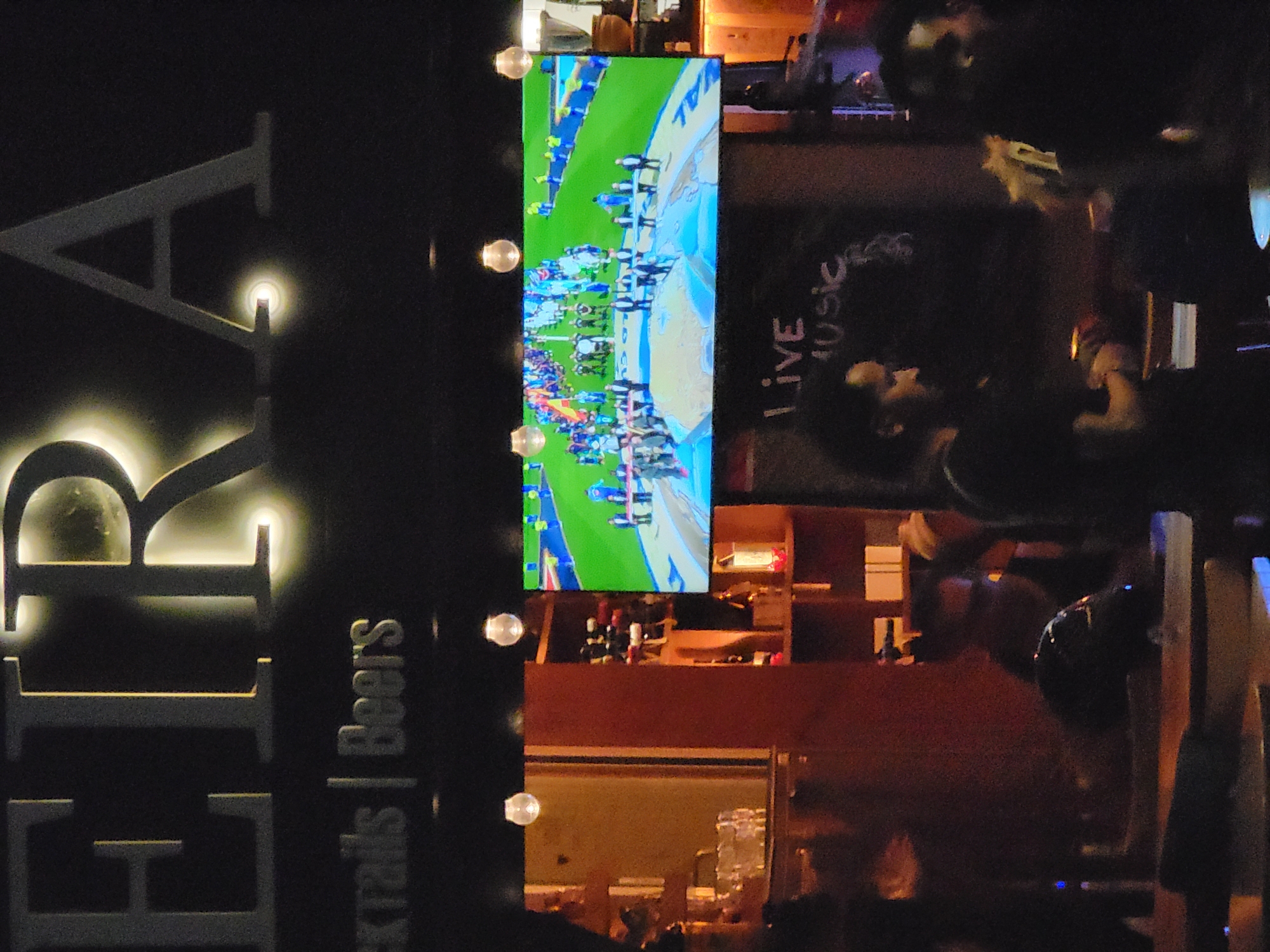 The image shows a dimly lit interior of a venue, possibly a bar or restaurant, with the word "EIRA" illuminated in large, glowing letters at the top. Below the main sign, smaller text reads "Cocktails | Beers". A large screen displays a brightly lit sports event, likely a soccer match, with players in uniforms on a green field. In the foreground, several people are visible, some in shadow, and others partially illuminated by the ambient light. A man in a black t-shirt is standing at what appears to be a counter or bar. The environment includes shelves with bottles, glasses, and decorative lighting. The overall impression is of a lively evening establishment. Given the prominent signage, it is likely located in Quarteira, Portugal.