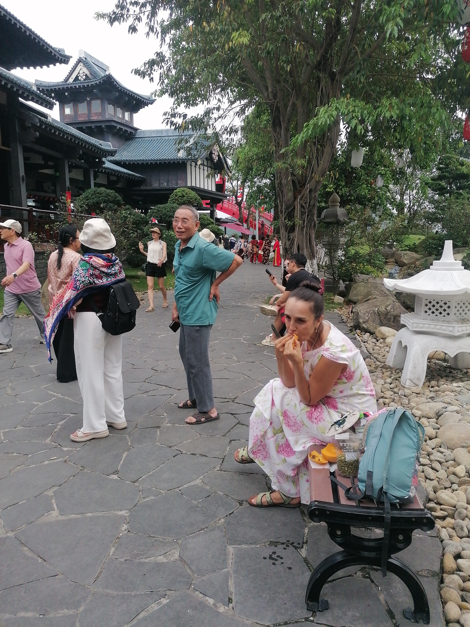 A woman is seated on a black bench in Hong Gai Ward, Vietnam. She is wearing a floral dress and sandals, and is eating a piece of fruit. A light blue backpack is leaning against the bench beside her. A container of seeds and a bottle are placed between her and the backpack.
Around her, there are various people walking along a stone pathway. Some are dressed in casual clothing, and appear to be tourists. An Asian-style building with multiple tiered roofs stands in the background to the left. To the right are trees, rocks, and a small white pagoda-like structure. A red bridge can be seen in the distance, partially obscured by the trees. The pathway is paved with irregular gray stones. The weather appears overcast.