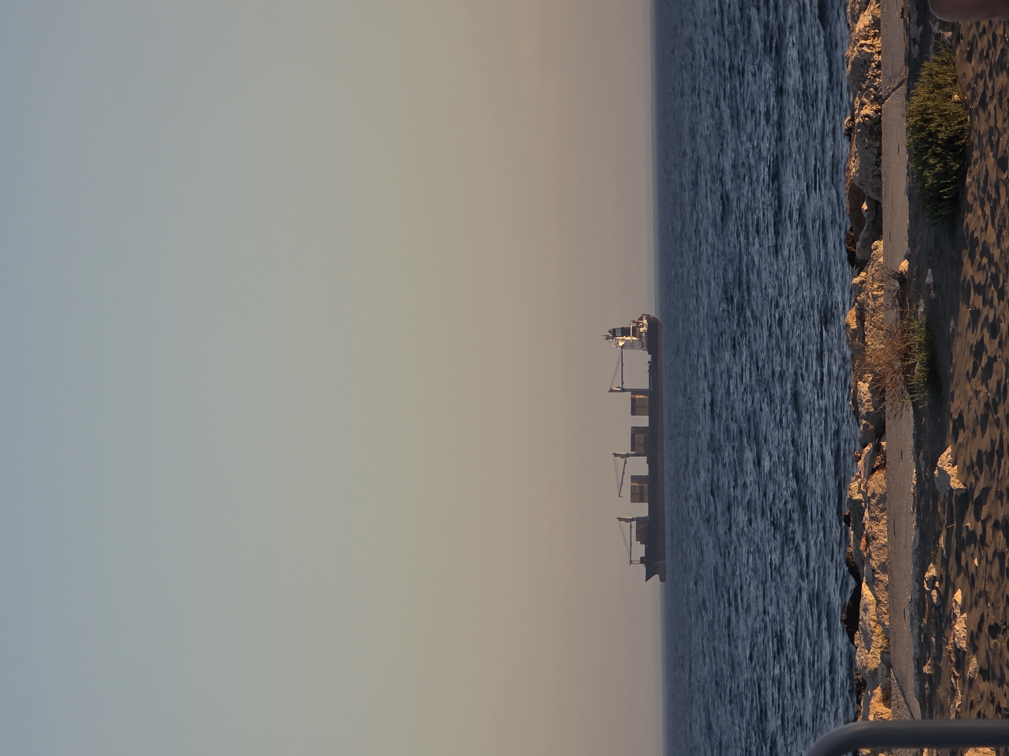 A cargo ship carrying containers is visible on the horizon in the blue sea under a clear, pale orange sky. The ship is dark-hulled and has multiple decks with stacked yellow containers. In the foreground, a rocky shoreline meets a sandy beach. Small green plants are growing on the sandy area. A portion of a light-colored metal railing is visible on the left edge of the frame. The image appears to be taken at dusk or dawn. The scene is likely from a coastal area in Málaga, Spain, given the Mediterranean Sea and typical shoreline features.