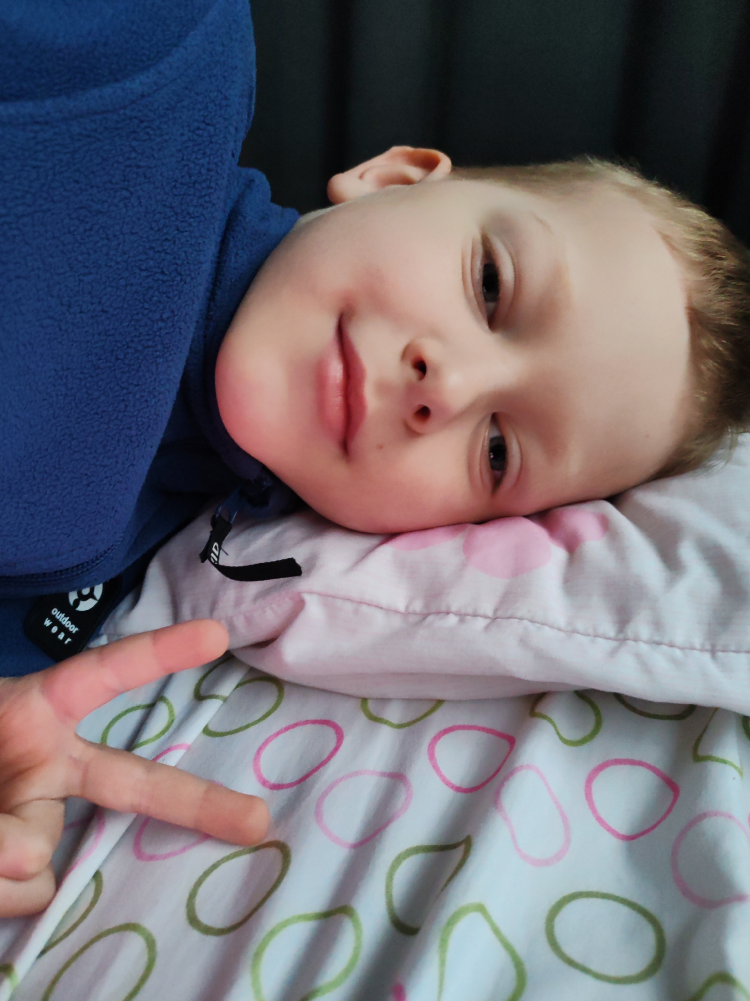A fair-skinned child lies on a white pillow with pink and green oval patterns on a bedsheet with the same pattern. The child is wearing a blue fleece jacket with a "outdoor wear" tag visible. The child is making a peace sign with their left hand. The background is a dark, blurry curtain. There is no clear indication of the location being the city of Chaikovsky, Russia.