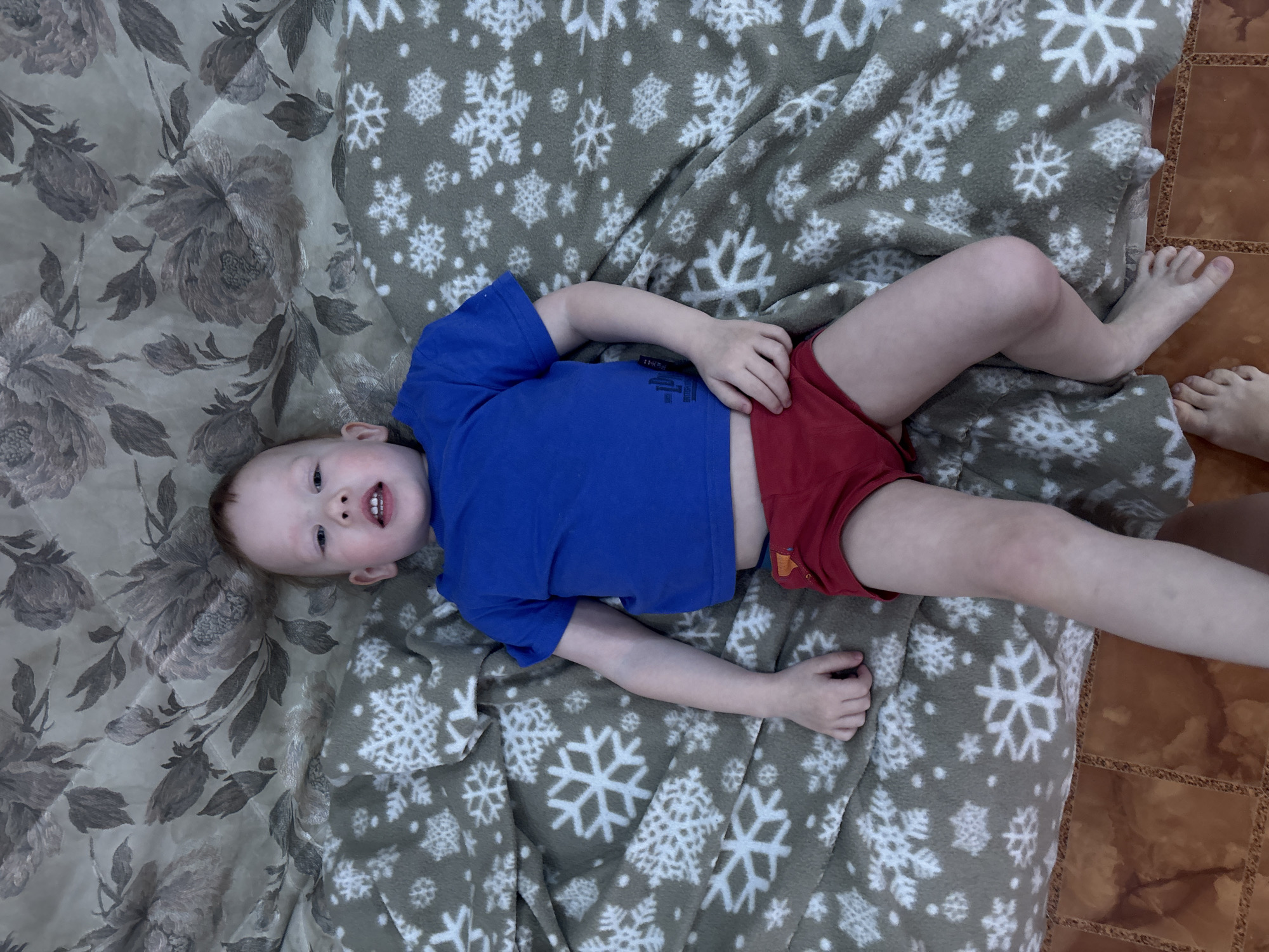 An overhead photograph shows a young child lying supine on a bed. The child, with fair skin and light brown hair, wears a blue short-sleeved t-shirt and red underwear. Their mouth is slightly open, revealing teeth, and eyes are directed towards the camera. The child is positioned on a rumpled grey fleece blanket featuring white snowflake patterns. Beneath this blanket, a lighter, quilted fabric with large, faded floral designs in shades of grey and brown is visible. The floor beneath the bed is covered with reddish-brown square tiles, each featuring a textured, braided border pattern. A portion of a bare human foot is visible in the lower left quadrant of the image. The specific location within the city of Ферма, Russia, is not visually determinable from the provided image.