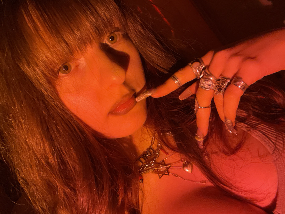 A woman with long brown hair and bangs poses in a dramatic red light. She wears multiple rings on her fingers with long, painted nails and several necklaces, including one with a Star of David. Her eyes are light colored. Her finger is touching her lips, and the overall effect is sultry and mysterious. The lighting adds a bold and theatrical quality to the portrait.