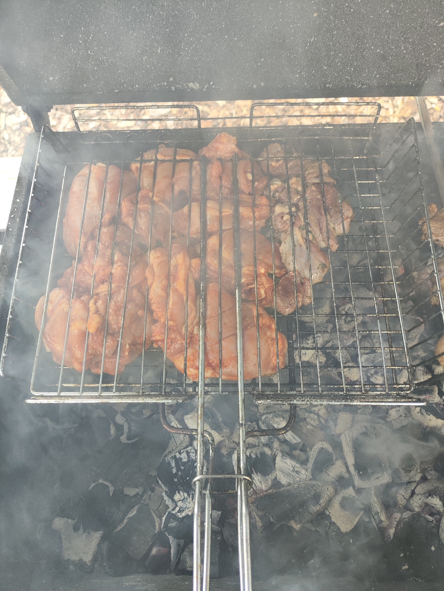This is a close-up, top-down shot of meat being grilled over hot coals in a black metal barbecue grill. The grill is filled with raw meat, some of which appears to be chicken thighs seasoned with a reddish spice mix, and other pieces that look like pork or beef strips. Smoke rises from the coals, obscuring some of the details of the food and the grill. The background of the image is blurred and appears to be an outdoor setting, possibly with leaves on the ground. There are no people or other animals visible in the image. The scene is set in the city of Старое Мартьяново, Russia.
