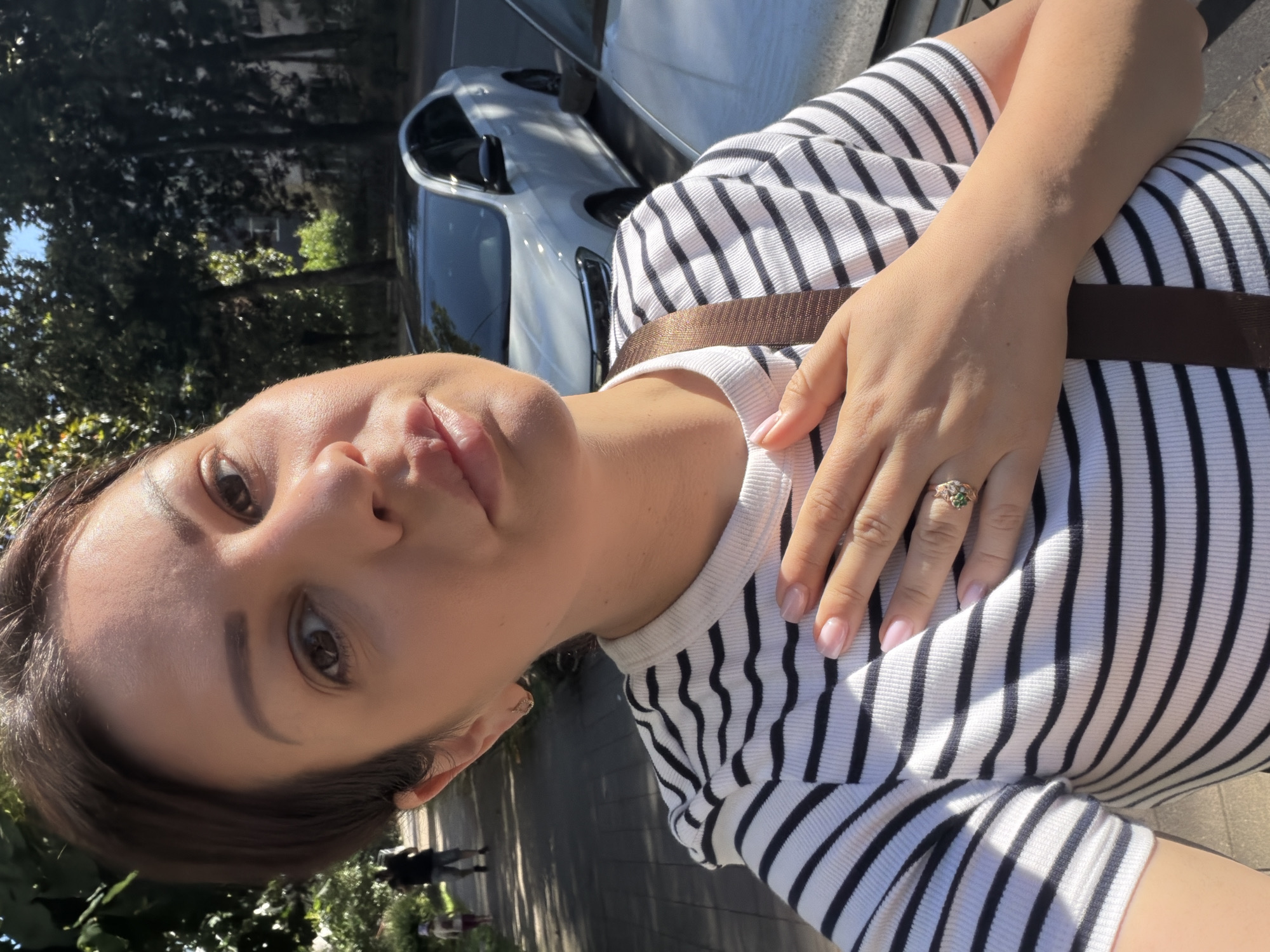 A woman with short, dark hair and fair skin takes a selfie outdoors on a sunny day. She is wearing a black and white striped t-shirt and a brown strap across her chest. Her right hand is placed on her chest, revealing a ring with a green stone and small white stones. In the background, a sidewalk is visible with two people walking in the distance. Trees line the sidewalk, and cars are parked on the street. The scene appears to be a residential street or park in Sochi, Russia.