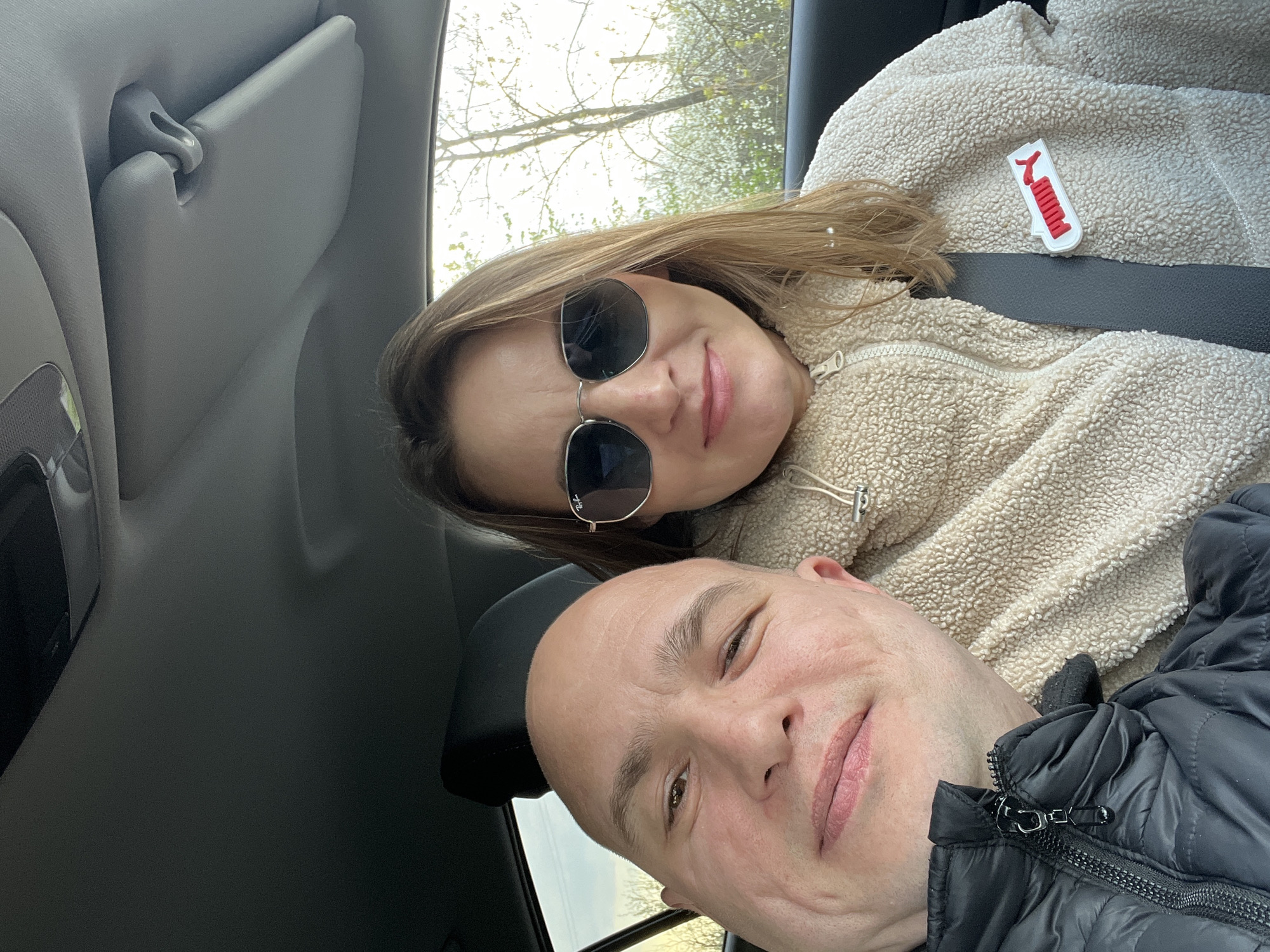 Two people are in a car, likely taking a selfie. The woman is wearing sunglasses and a tan, fleece jacket with a Puma logo. The man has a shaved head and is wearing a black jacket. The car's interior is visible, including the sun visor and a glimpse of trees through the window. The location is Unknown, Unknown.