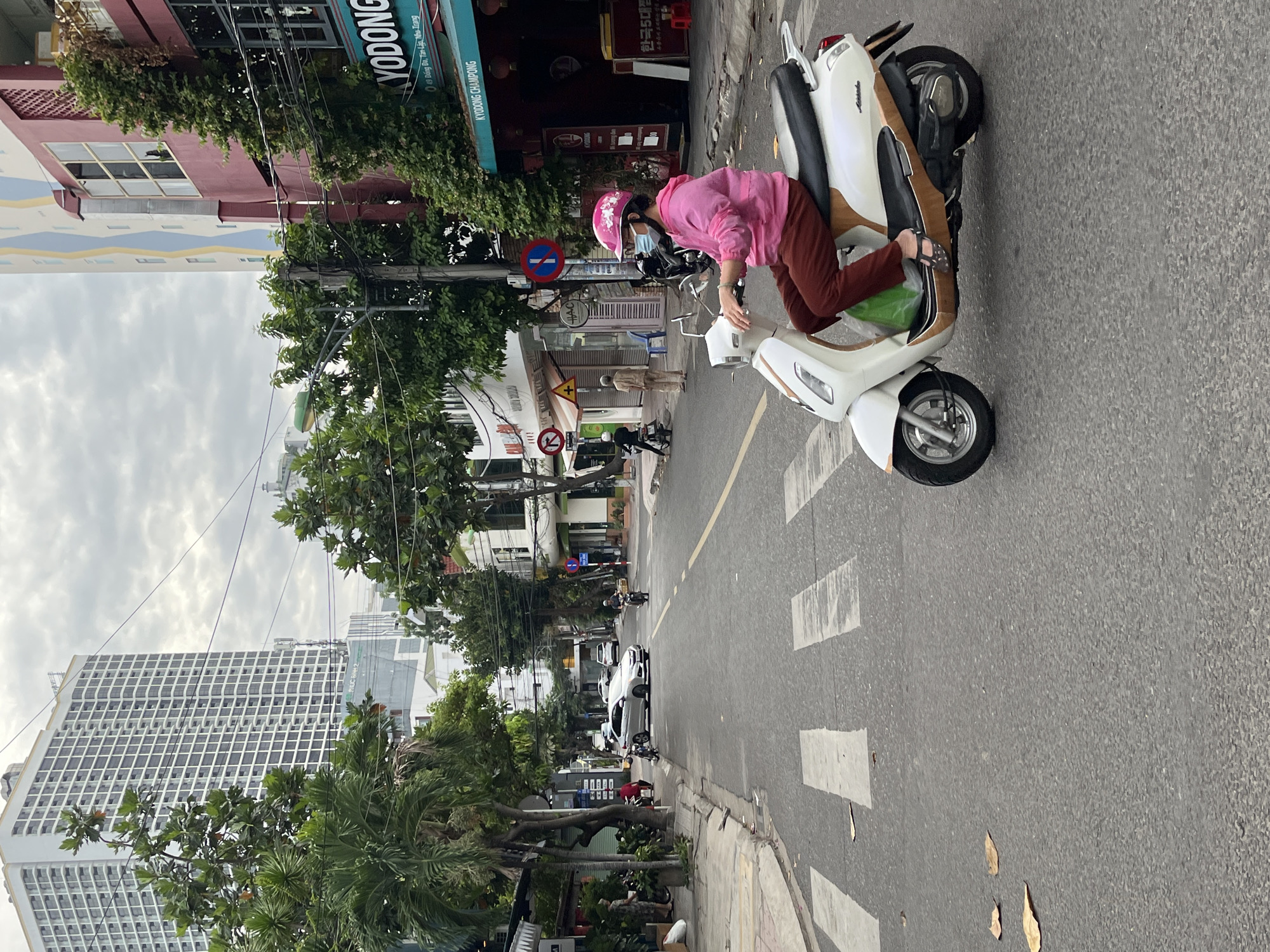 In Nha Trang, Vietnam, a woman wearing a pink hooded jacket, brown pants, and a pink helmet with a white flower design rides a white scooter. She is also wearing a blue face mask. The scooter has brown accents on its body. The woman is on a street with a crosswalk in the foreground and is heading away from the viewer. Trees and buildings line the street. A white car is parked on the left side of the street, and another car and scooter are visible further down the road. Overhead power lines are present. In the background, a tall white building with many windows stands out, and a red building with the word "KYODONG" in green lettering is visible. The sky is overcast.