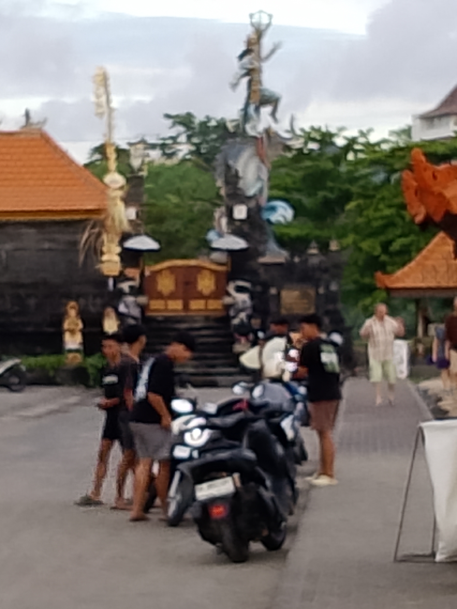 The image depicts a street scene in Kediri, Indonesia, likely during the late afternoon or early evening given the overcast sky and muted lighting. In the foreground, several motorcycles are parked. A group of young men, dressed casually in t-shirts and shorts, are standing and conversing near the motorcycles. One man is wearing a black t-shirt with a visible white graphic on the back.
Behind the group, a pathway leads to a building with traditional Balinese architectural elements, characterized by ornate carvings and a distinctive red-tiled roof. A large, elaborate statue of a figure, possibly from Hindu mythology, stands prominently at the entrance to the building, surrounded by lush green trees. Further down the pathway, a man in a checkered shirt and light-colored shorts walks away from the camera, his posture suggesting he might be gesturing. Another person is partially visible walking on the path. The overall atmosphere appears relaxed and casual, characteristic of a local gathering place or a tourist area. There is no visible text in the image.
