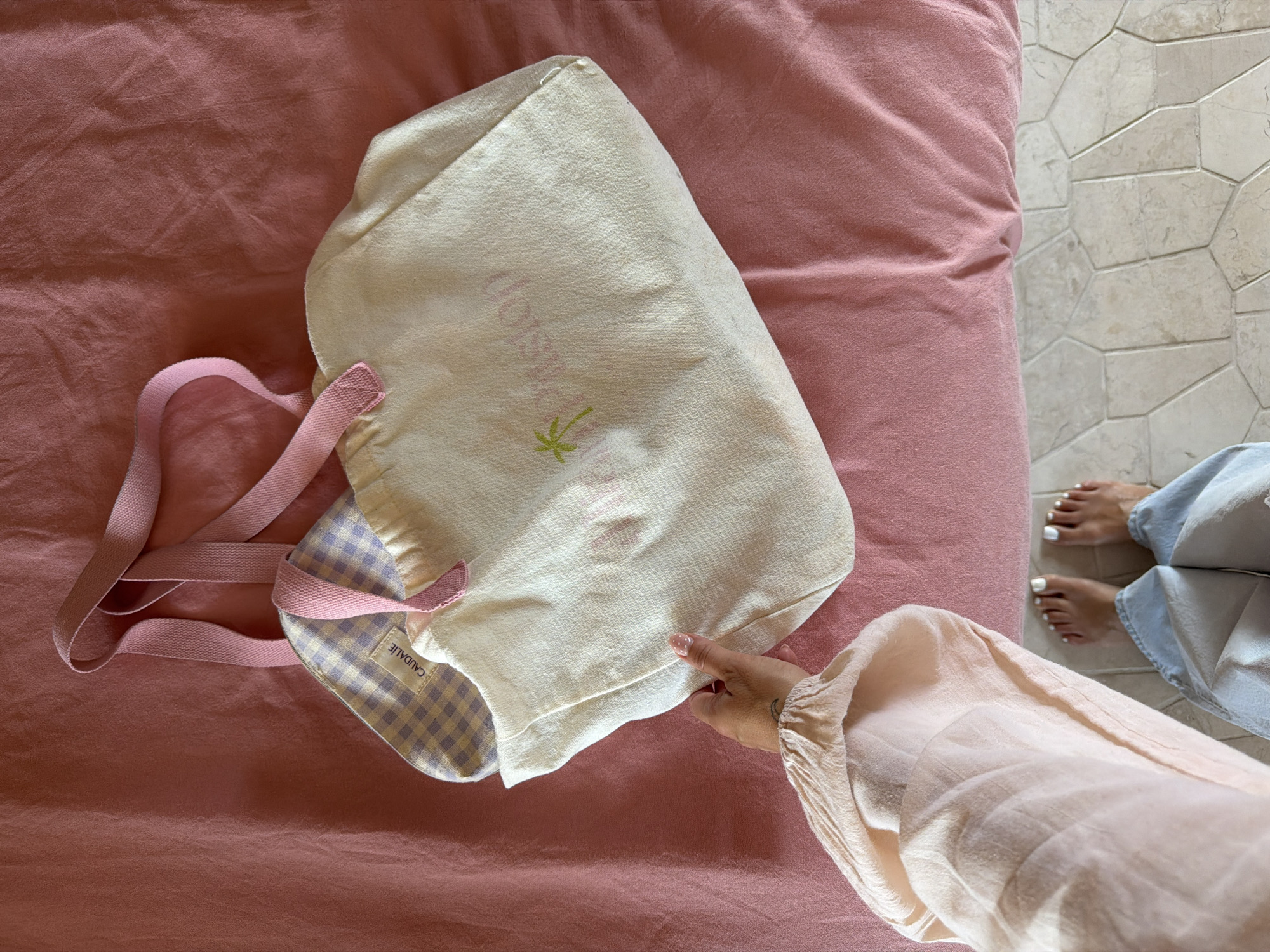 A top-down shot displays a person's hand holding a canvas tote bag on a pink linen surface. The tote bag is off-white with pink straps and a pastel gingham interior. A palm tree graphic and pink text are visible on the bag. The person's forearm, wearing a pale peach linen sleeve, is in the lower left corner. Their bare feet with white toenail polish are visible in the bottom center, standing on a hexagonal tiled floor. The scene is indoors, in a room located in the city of Mengwi, Indonesia.