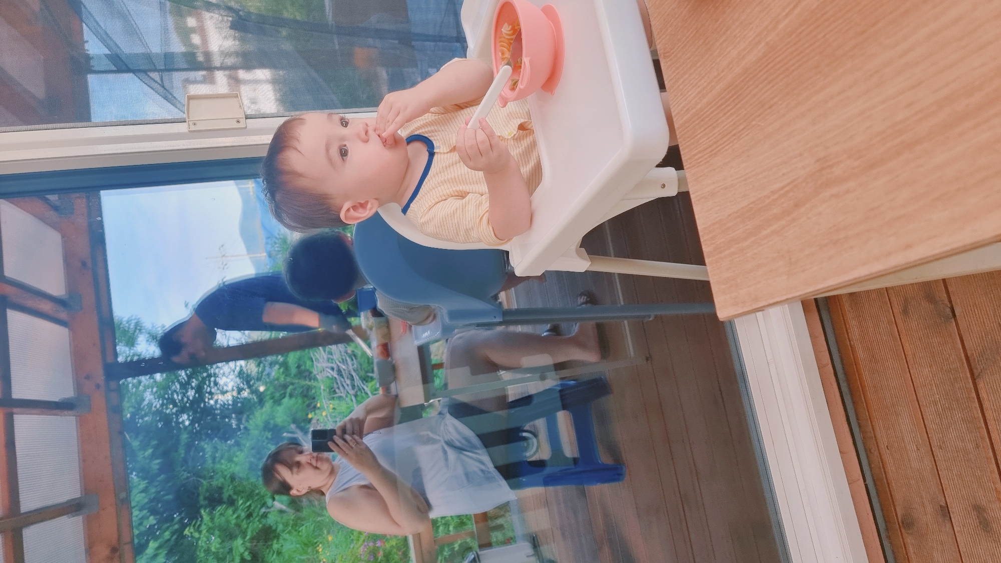 A baby sits in a white highchair eating from a pink bowl. The child is fair-skinned, with brown hair and eyes, wearing a yellow and white striped shirt with blue trim. The baby holds a white spoon in one hand and food in the other, with a focused expression. The highchair is positioned indoors next to a window overlooking an outdoor scene.
Visible through the window are two adults: one wearing a blue shirt bending over and the other holding a smartphone in her hands. The reflection of the outdoor scene includes greenery, trees, and a blue sky suggesting a rural or natural environment. The location is likely Dunnae-myeon, South Korea, as indicated by the surrounding environment and architecture visible in the reflection, and the natural light.
The scene depicts an everyday domestic moment.