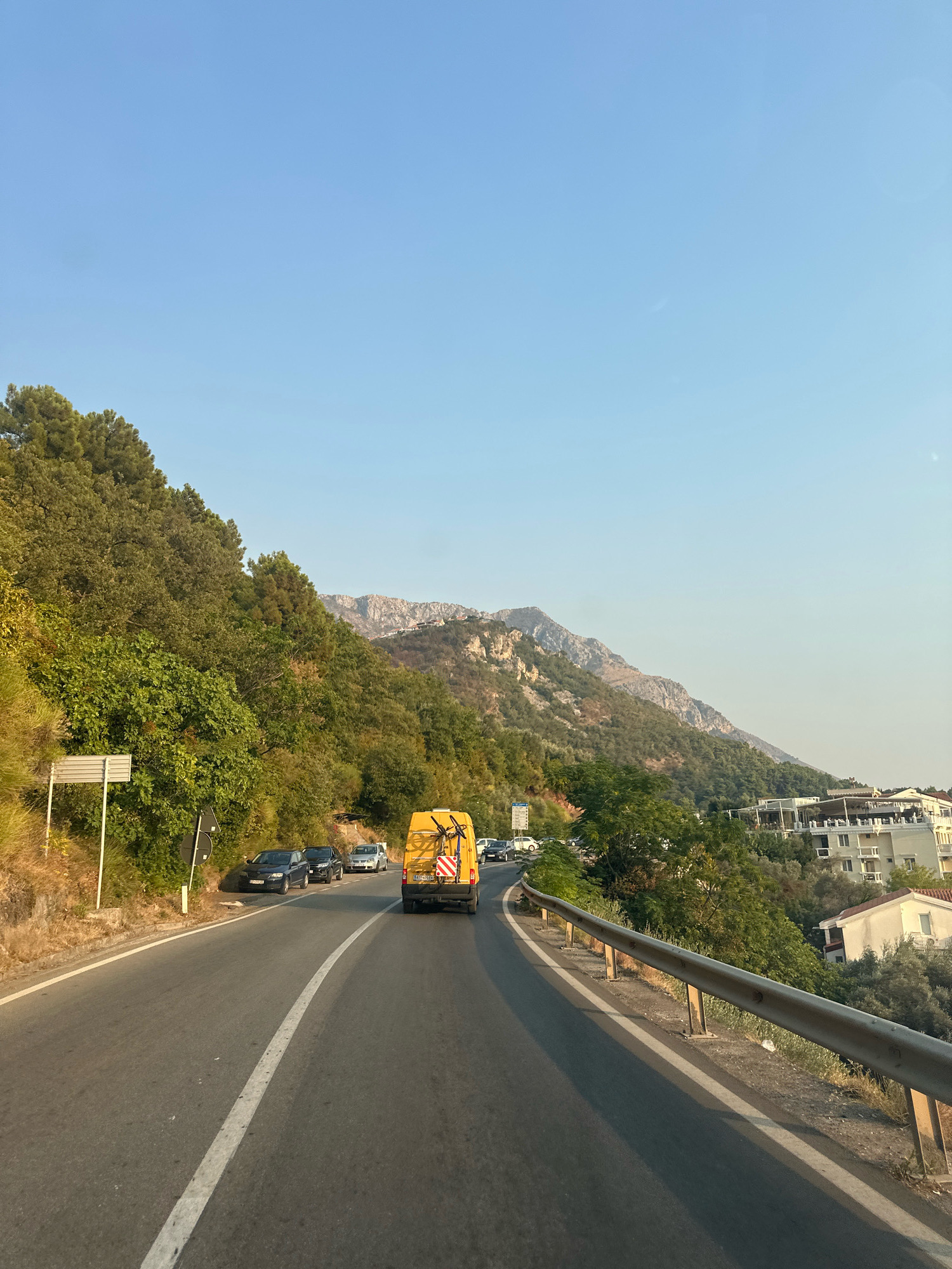 The image depicts a scenic view from a vehicle traveling along a coastal road near Sveti Stefan, Montenegro. The road curves to the left, with a steep, tree-covered mountainside on the left and a drop-off to the right, protected by a metal guardrail. Several cars are parked and driving on the road. A yellow van, possibly a service vehicle, is in the foreground, also traveling along the road. In the distance, buildings and a more rugged mountain range are visible against a clear blue sky. The overall impression is one of a sun-drenched, picturesque drive through a Mediterranean landscape.