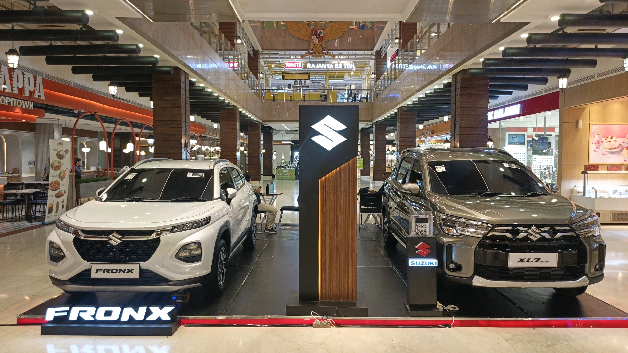 Two Suzuki cars, a white Fronx on the left and a grey XL7 New Zeta on the right, are displayed on elevated platforms in a shopping mall. A large, black pillar with the Suzuki logo and wooden accents stands between the cars. The background shows a multi-level mall interior with shops, lighting fixtures, and decorative elements, including an eagle emblem on a higher level. The location is identified as City of Medan, Indonesia. There are no discernible interactions between people and the cars, and the scene appears to be a static car display.