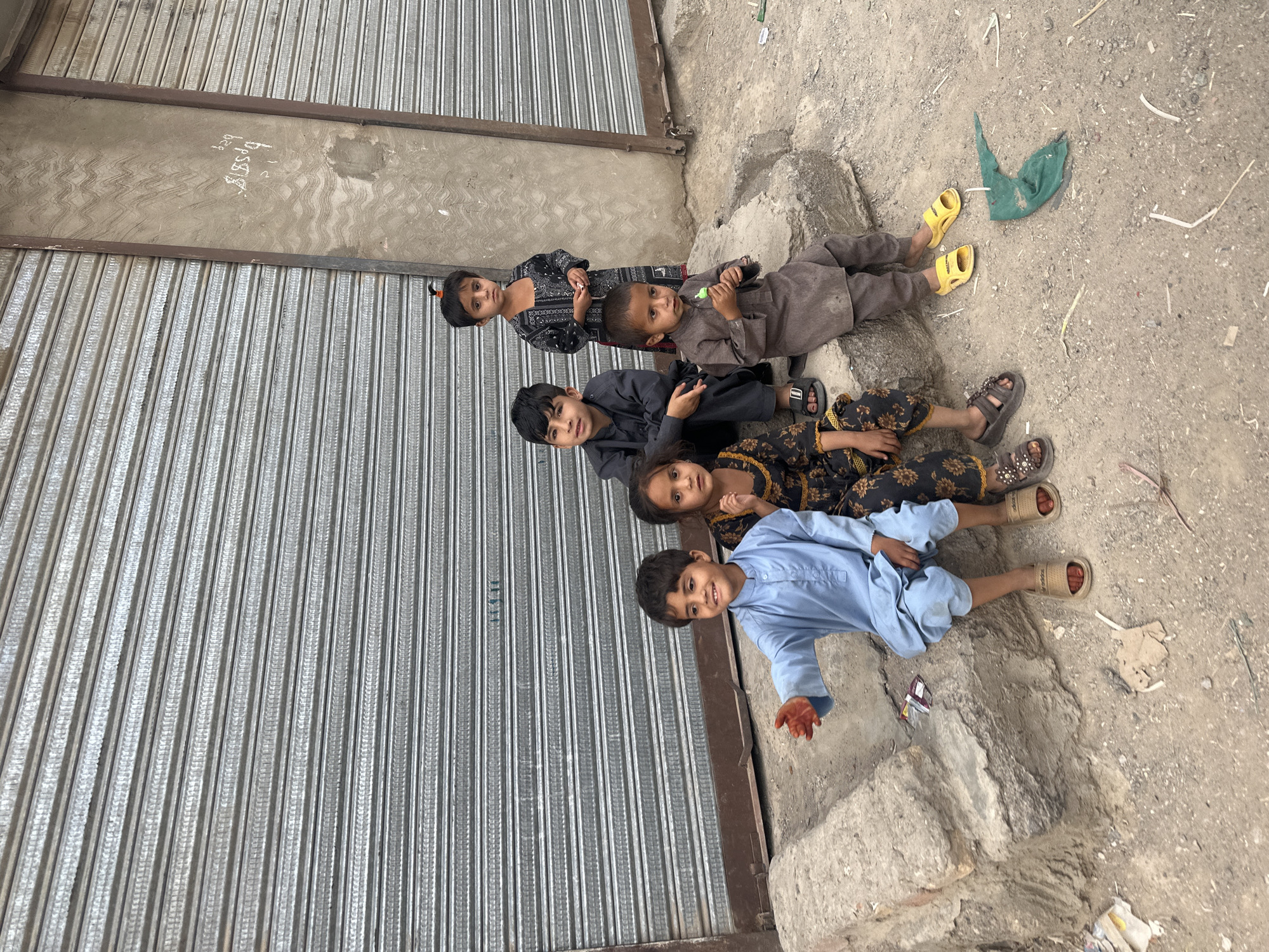 Five children lie in a row on the ground next to a closed metal shutter in Quetta City Tehsil, Pakistan. The shutter appears to be part of a building or store. The ground is cracked concrete and dirt. A small amount of litter is scattered nearby.
The children are dressed in varying styles of clothing: a light blue outfit, a floral patterned outfit, a black outfit, a brown outfit, and a black patterned outfit. They are all wearing sandals or slippers.
The children are positioned in a line, with the child in the light blue outfit closest to the viewer and the child in the black patterned outfit furthest away. They appear to be posing for a photograph.