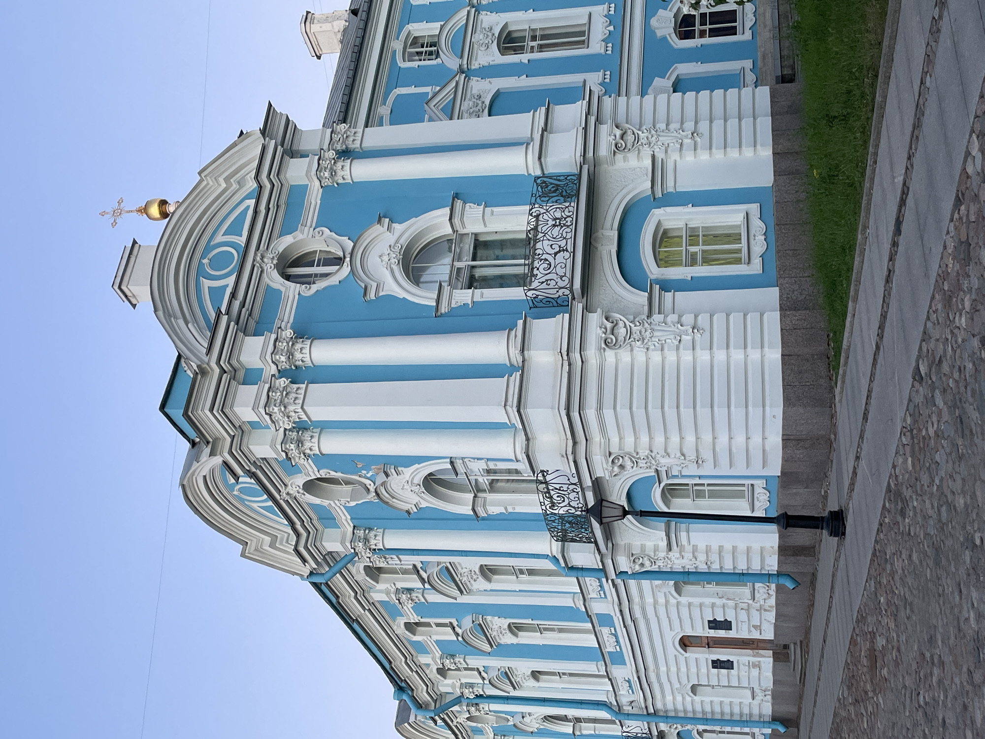 The image shows the Smolny Cathedral complex in Saint Petersburg, Russia, specifically the southwestern facade of the Smolny Convent's bell tower. The building is painted a striking blue and white, with ornate architectural details. The facade features multiple arched windows, some with decorative balconies, and prominent white columns with Corinthian capitals. A golden dome topped with a cross is visible on the roofline. The foreground shows a cobblestone street and a sidewalk, with a black lamppost standing near the building. The sky is clear and pale blue, indicating it might be early morning or late evening. There are no people or vehicles present in the image.