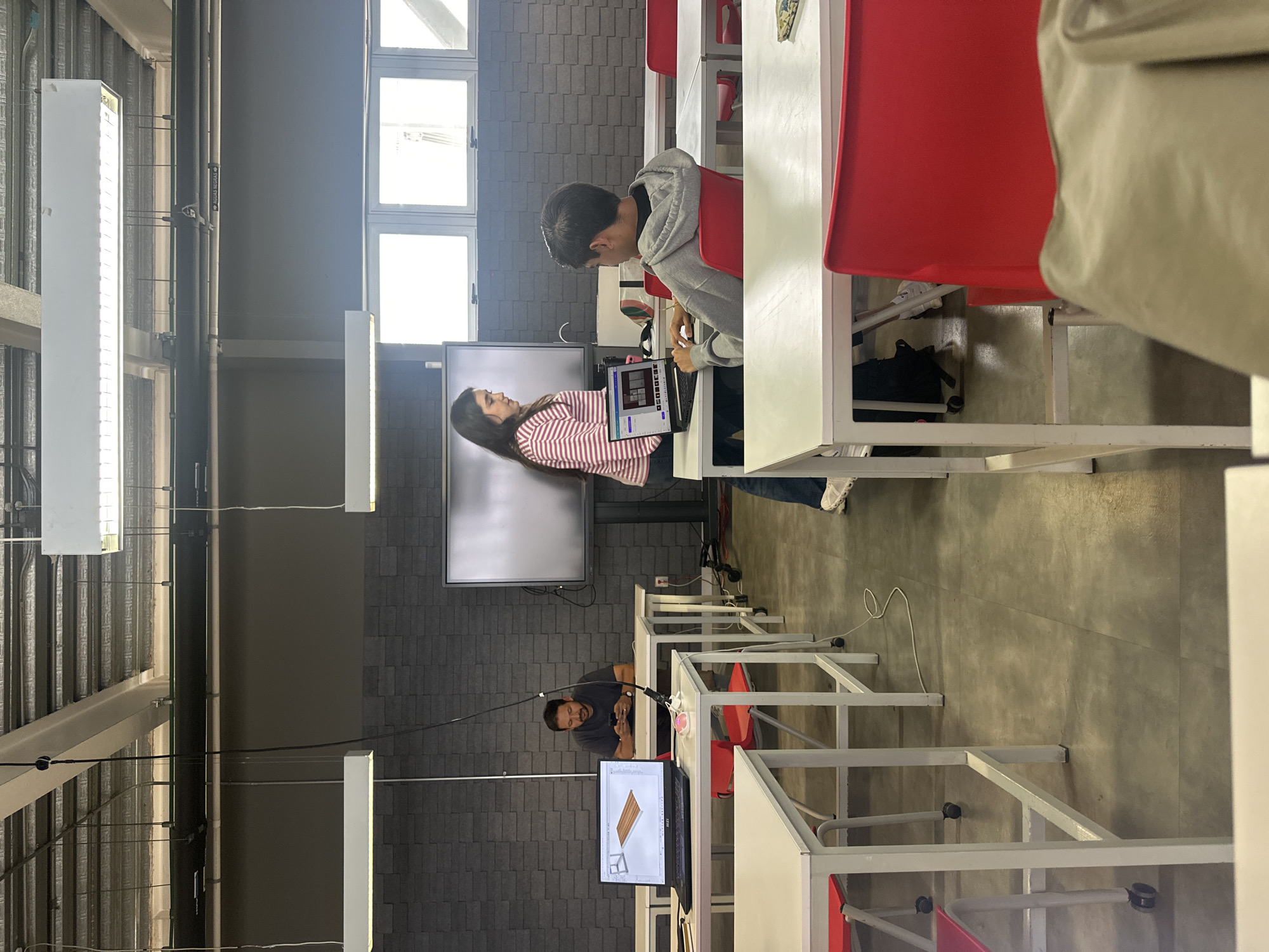 A classroom scene features two students, a male and a female, engaged in a collaborative project displayed on a laptop. The male student is seated, while the female student stands beside him, both focusing on the screen. A larger display screen is mounted on the wall behind them. Desks with red chairs are arranged in rows throughout the room. In the background, a male individual is visible using his phone near a monitor. The room is illuminated by fluorescent lights hanging from the ceiling. Windows provide natural light, and the walls are constructed with gray brick or tile. The exact location in Mexico City cannot be determined from the image alone.