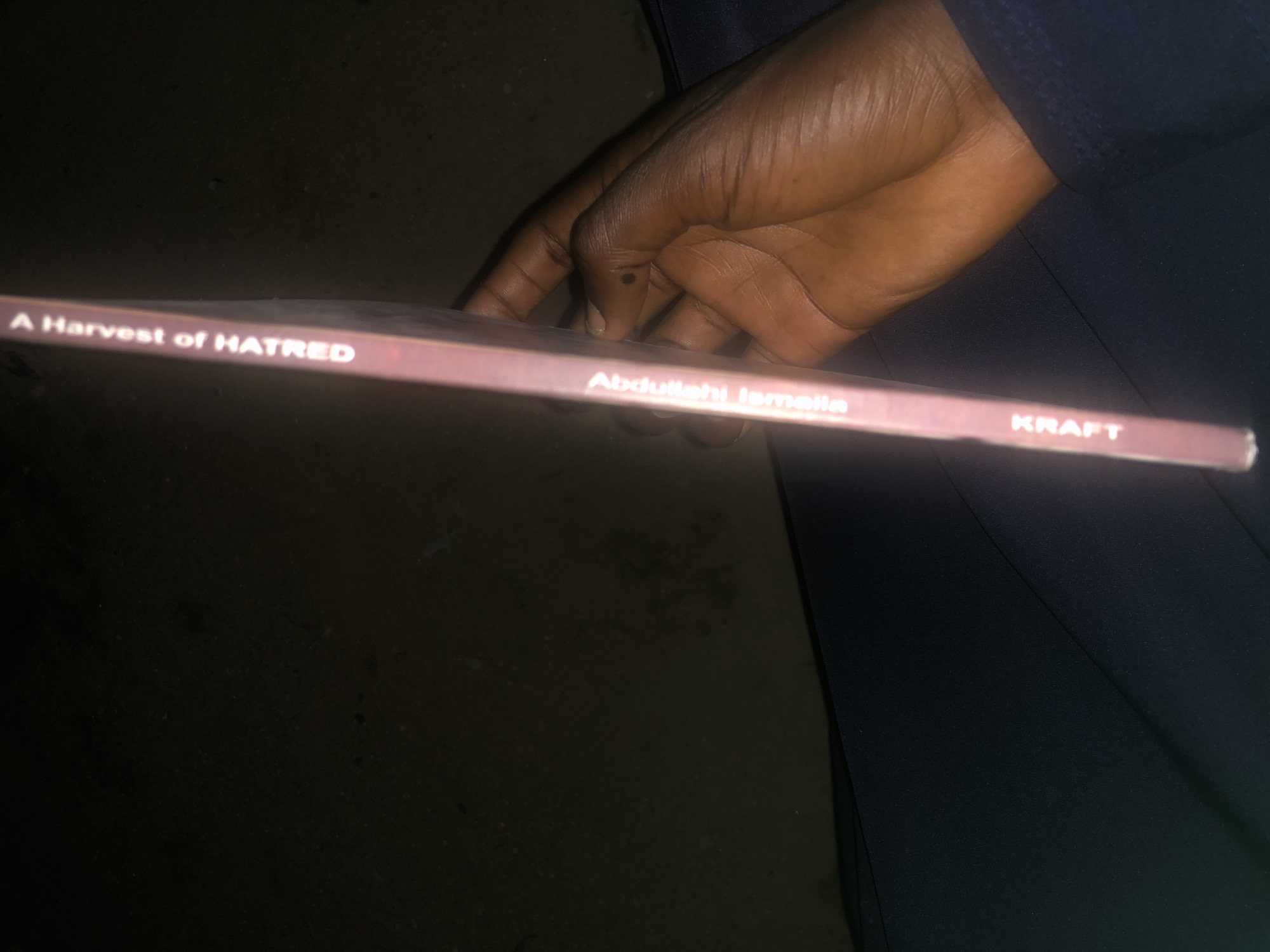 The image shows the spine of a book titled "A Harvest of Hatred" by Abdullahi Ismaila, published by KRAFT. A person's hand is holding the book. The background is dark and appears to be an indoor setting. There are no discernible landmarks or environmental features to place the image specifically within Jalingo, Nigeria.