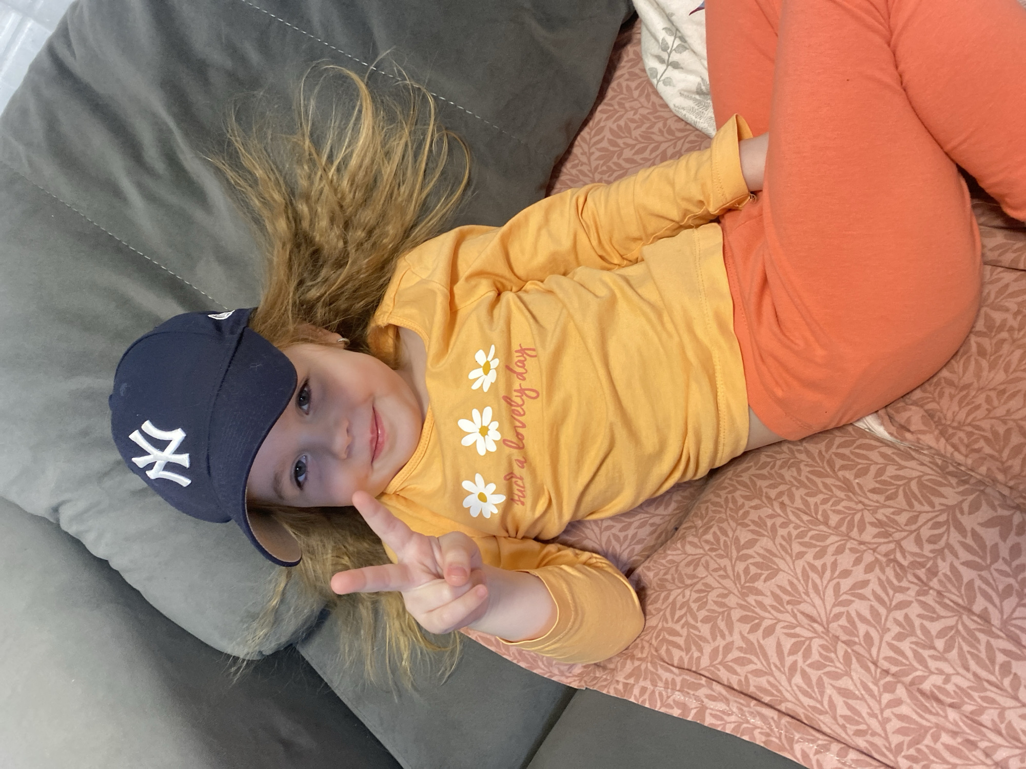 A young child with blonde hair is lying on a patterned pink blanket, dressed in a long-sleeved yellow shirt with daisy graphics and orange leggings. The child is wearing a dark blue baseball cap with a white "NY" logo. The child is giving a peace sign with their right hand. A gray pillow is positioned to the left of the child. There is no verifiable information within the image to confirm a location in Izhevsk, Russia.