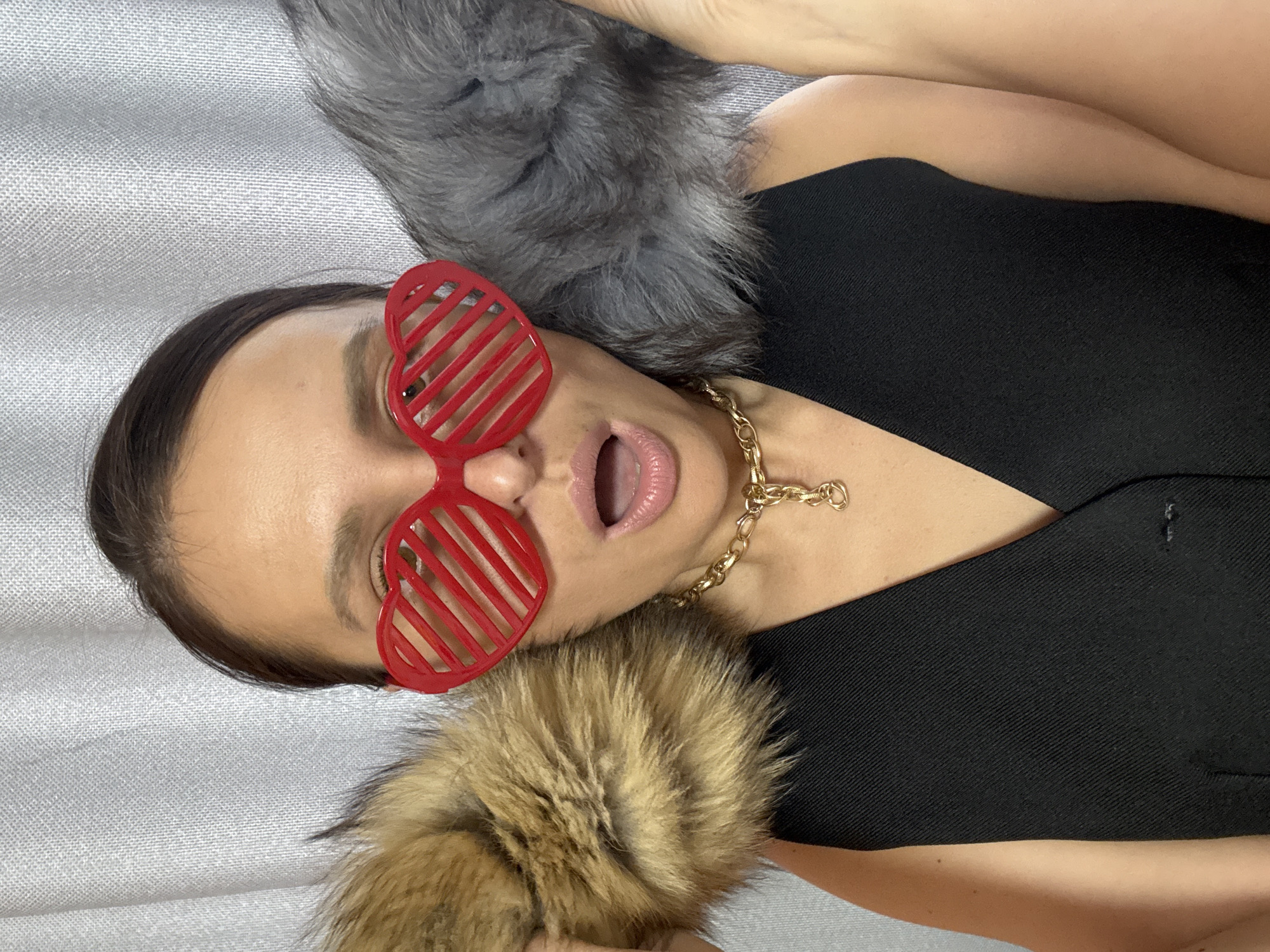 A close-up, eye-level image captures a woman lying down or reclining, viewed from her chest upwards, with her head tilted. She has dark hair pulled back and wears striking, red, heart-shaped novelty sunglasses with horizontal slats. Her mouth is open, revealing her tongue, suggesting a playful, expressive, or surprised demeanor. Around her neck is a prominent gold-colored chain necklace with an indistinct small pendant. She is dressed in a black garment, possibly a top or vest, with a structured neckline.
Two fluffy fur items are visible: one grey-silver and one brown-tan, appearing either as part of her attire (like a collar or trim) or as separate soft accessories she is interacting with. The background is a textured, light-grey fabric, indicative of an indoor setting such as a sofa or bed. The lighting is even, and there are no discernible location cues to specify Chaikovsky, Russia, or any particular time of day. No text is visible. The overall mood is casual and lighthearted.