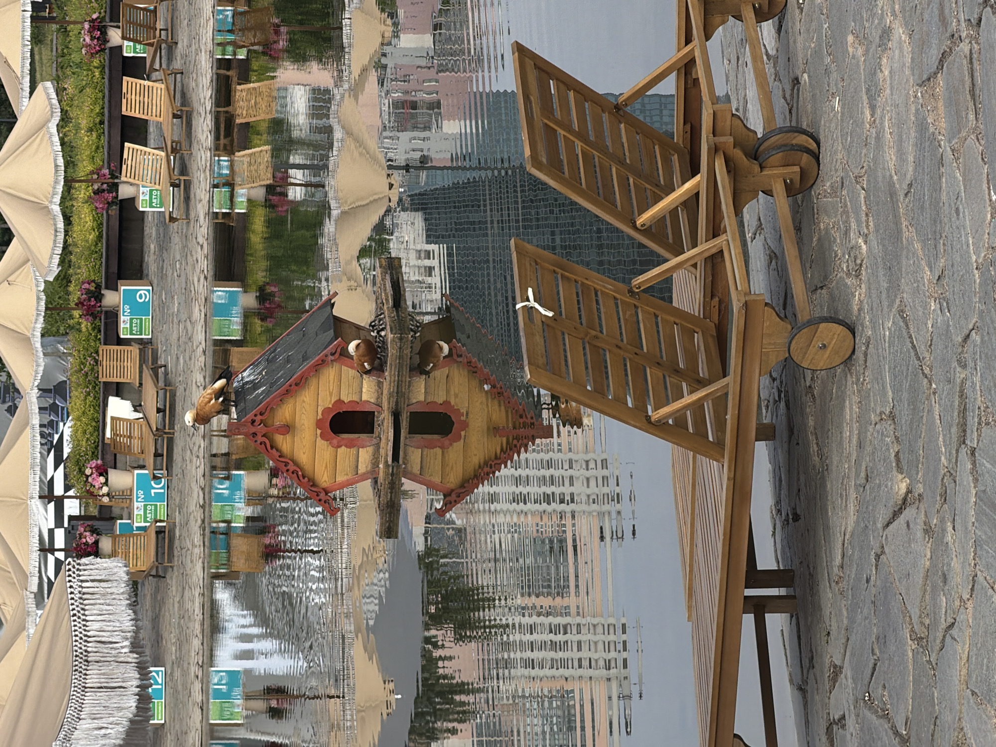 The image captures a serene waterside scene in Khimki, Russia, likely at a park or resort. In the foreground, two wooden chaise lounges with wheels are positioned on a stone-paved area, facing a body of water. The lounges are empty. The water reflects buildings and umbrellas, creating a distorted, impressionistic view. In the middle ground, a small, whimsical wooden house, seemingly a birdhouse or decorative feature, floats on the water. A ruddy shelduck is perched on top of this structure, and a few ducklings are huddled on the platform. In the background, several large, beige umbrellas with fringed edges offer shade, and lounge chairs are visible on the paved area behind them. Floral arrangements in pots add a touch of color to the scene. The overall impression is one of a calm, recreational environment.