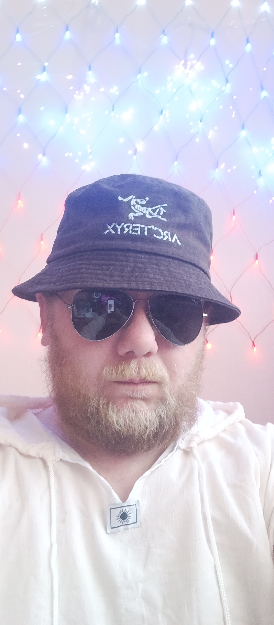 A person with a beard and sunglasses is wearing a dark bucket hat with a logo, and a white hooded shirt with a tag featuring a sun symbol. The background consists of string lights, alternating between red and blue. The location is the city of Unknown, Unknown.