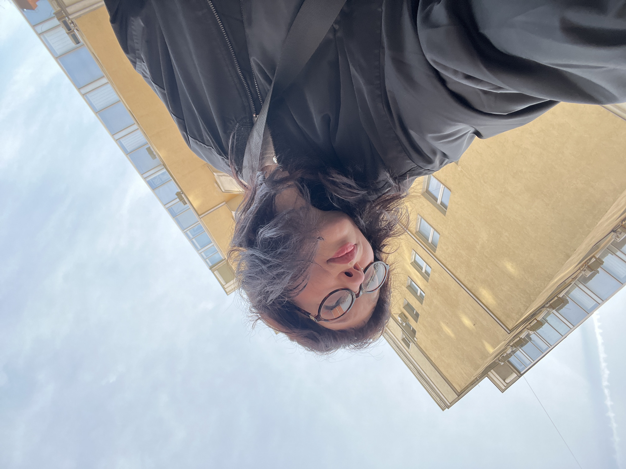 A woman wearing glasses and a black jacket is viewed from a low angle, looking downwards. In the background, two buildings are visible. One building is yellow and appears to be a residential apartment building, with several windows. The second building has an angular facade with rows of windows. The sky is visible behind the buildings, with a vapor trail cutting across the sky.
While precise location information within Kazan, Russia, is not directly discernible from the image, the architectural style of the building suggests it is an urban setting.