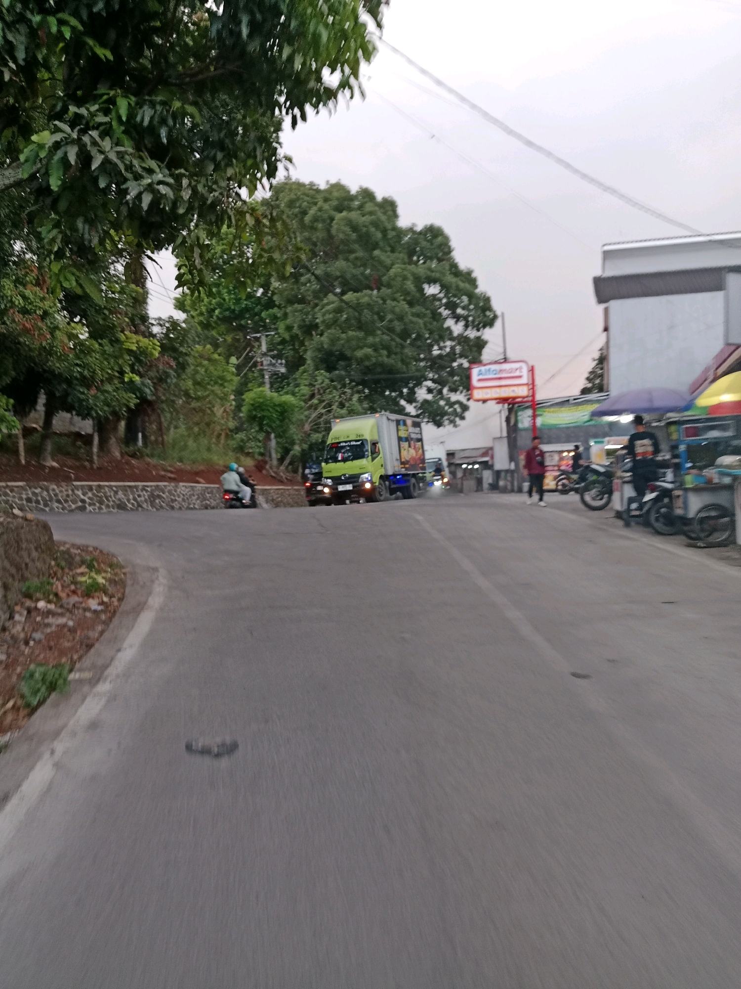 The image depicts a street scene in Cilaku, Indonesia. The road curves to the left in the foreground, with a stone retaining wall and vegetation on the left side. A light green truck is driving on the road, and two people are on a motorcycle behind it. On the right side of the road, there are several shops and street vendors. One shop has an "Alfamart" sign. There are also parked motorcycles and people standing around. The sky is overcast.