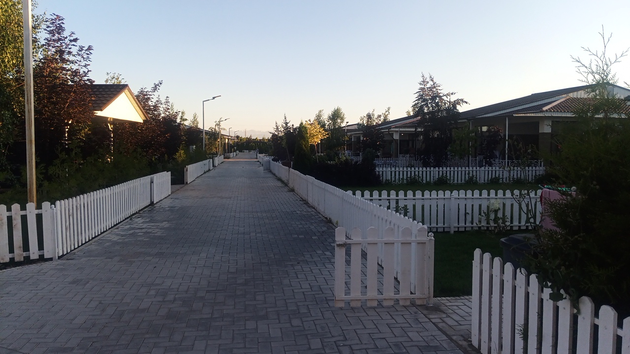 A paved walkway lined with white picket fences stretches into the distance, flanked by houses and trees under a clear sky. The walkway is made of grey rectangular pavers laid in a herringbone pattern. On the left side, a white picket fence follows the walkway, with some dark trees and a building with a triangular roof visible behind it. On the right side, another white picket fence runs parallel to the walkway, with the eaves of low-rise buildings with dark roofs visible behind it. The buildings appear to be modern homes or guest houses, with covered patios and large windows. The environment suggests a suburban or resort setting, possibly in a warm climate given the clear sky and greenery. The scene is devoid of people or any discernable activity. The lighting indicates it is either early morning or late afternoon, with the sun casting a warm glow on the scene. The location is identified as Choktal, Kyrgyzstan.