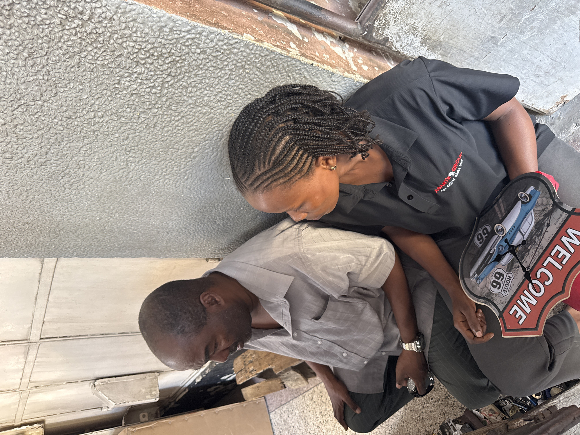 Two individuals, an adult male and an adult female, are seated on the ground next to a textured, grey wall. The male, on the left, is wearing a light grey button-down shirt and dark pants. He has a watch on his left wrist. The female, on the right, is wearing a black polo shirt with a logo that reads "ASANTE GROUP" and "we deliver what's best". Her hair is styled in cornrows. Between them, resting on their laps, is a decorative clock shaped like a red arrow sign with "WELCOME" written in white text and an image of a classic car with "ROUTE 66" on it. The environment appears to be an outdoor setting, possibly an alleyway or a covered walkway, with a rough concrete floor and a weathered metal door or panel in the background to the left. The image suggests a casual, everyday moment in Dar es Salaam, Tanzania, perhaps near a commercial area or a residential building.