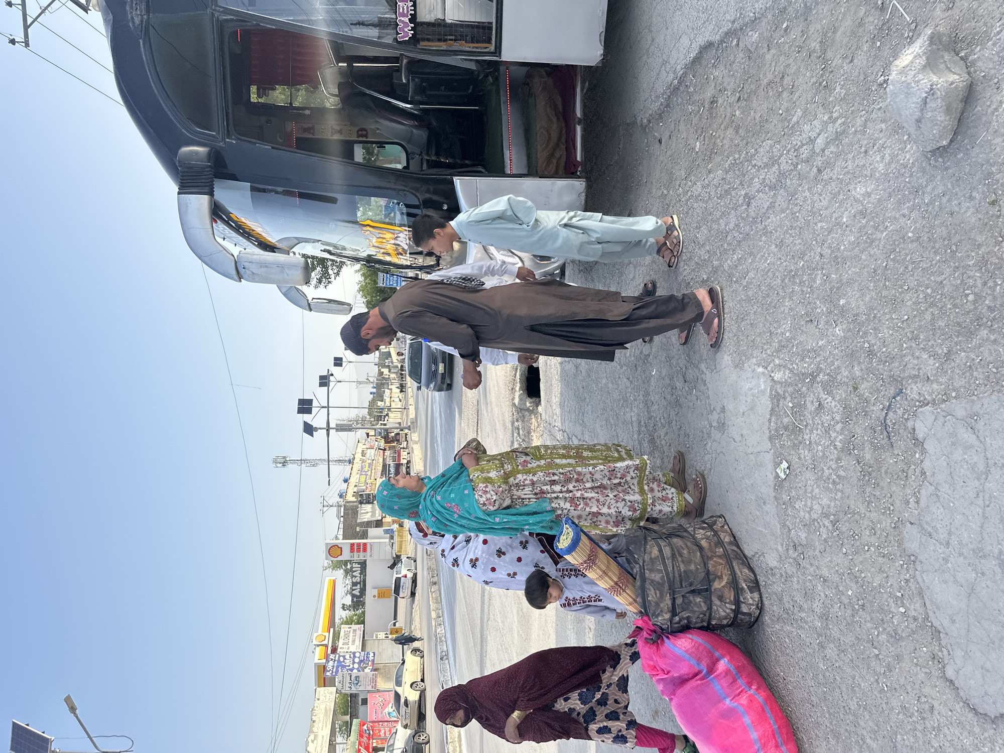 This media file captures an outdoor scene at a roadside bus stop or waiting area in Quetta, Pakistan, during clear daylight hours.
The primary subjects are a group of people and a large bus. A man wearing a dark brown shalwar kameez and a cap stands near the open door of a dark-colored bus, seemingly looking downwards. Beside him, a young boy in a light blue shalwar kameez also faces the bus. Further to the left, two women and a child are visible. One woman wears a floral printed dress with a turquoise headscarf, and the other wears a maroon headscarf over a patterned dress. They are accompanied by luggage, including a large dark bag and a bright pink sack, and appear to be waiting or in the process of boarding/disembarking the bus.
The setting is an urban or semi-urban environment. The ground is a mix of asphalt and dirt. In the background, a street with several cars, power poles, and commercial buildings can be seen. A prominent feature is a Shell petrol station sign displaying "AL SAIF" and fuel prices (e.g., 302.93, 299.98, 280.00). Other smaller signs and billboards are also visible, including one that reads "OIL CHANGE." The sky is clear and bright, indicating sunny weather.
The activity suggests a travel scenario, with people gathered around a bus, possibly preparing for a journey. The clothing, the bus type, and the scripts on the signs (primarily Urdu/Arabic) are characteristic of the region. A sticker with the text "MAA" is visible inside the bus.