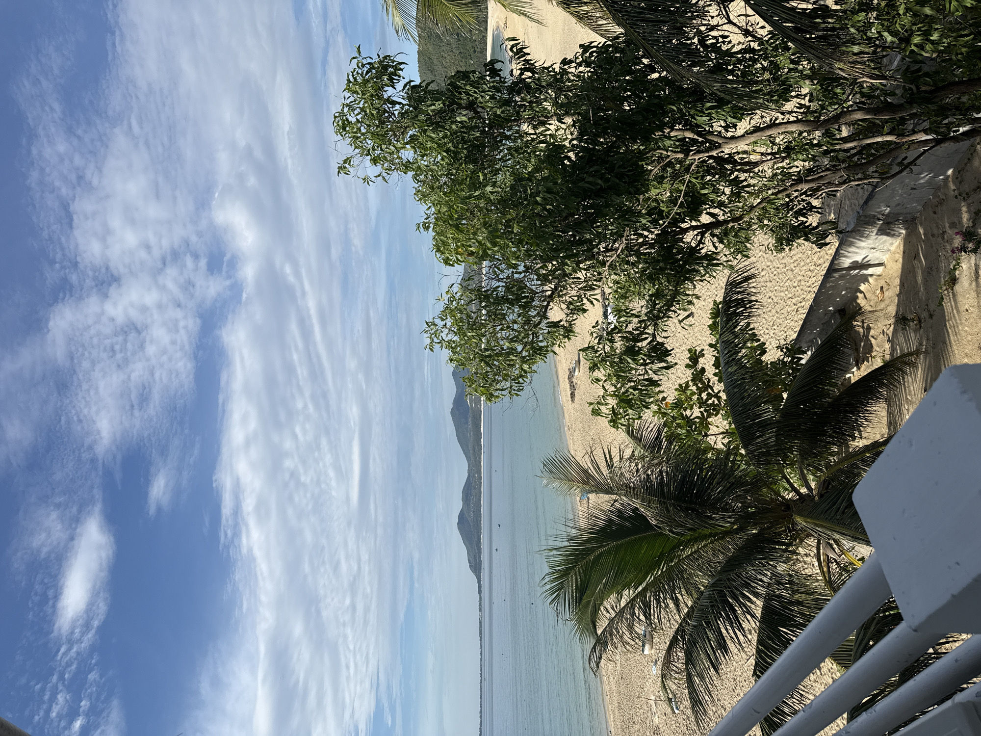 A wide-angle, eye-level shot captures a tranquil beach scene under a partly cloudy sky. The foreground features a white railing, with palm fronds and dense green foliage partially obscuring the view of the sandy beach. The ocean, a calm expanse of light blue water, stretches to the horizon, meeting a distant line of tree-covered mountains. A few small boats are scattered across the water. The sand is light brown and appears mostly smooth, with some evidence of gentle waves lapping at the shore. The sky is a vibrant blue, dotted with wispy white clouds.