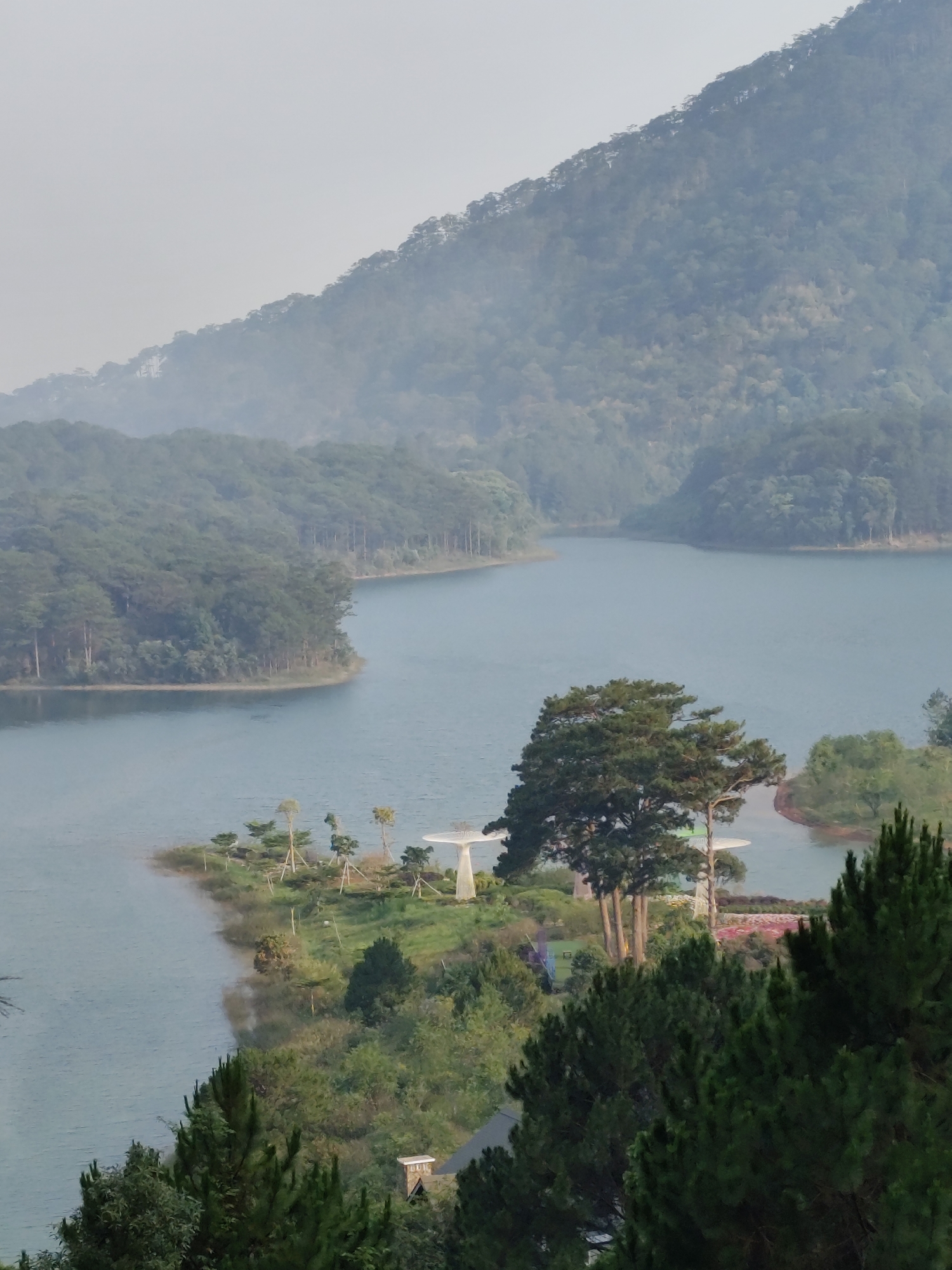 The image presents a scenic view of Tuyen Lam Lake in Da Lat, Vietnam, characterized by a misty atmosphere. The foreground features a peninsula covered with lush greenery, including pine trees and manicured gardens. Two white, mushroom-shaped structures are visible on the peninsula. The lake extends into the midground, winding between tree-covered hills. In the background, a mountain range rises into the misty sky, its slopes densely forested. The overall color palette is dominated by shades of green and blue, contributing to a tranquil and serene ambiance.