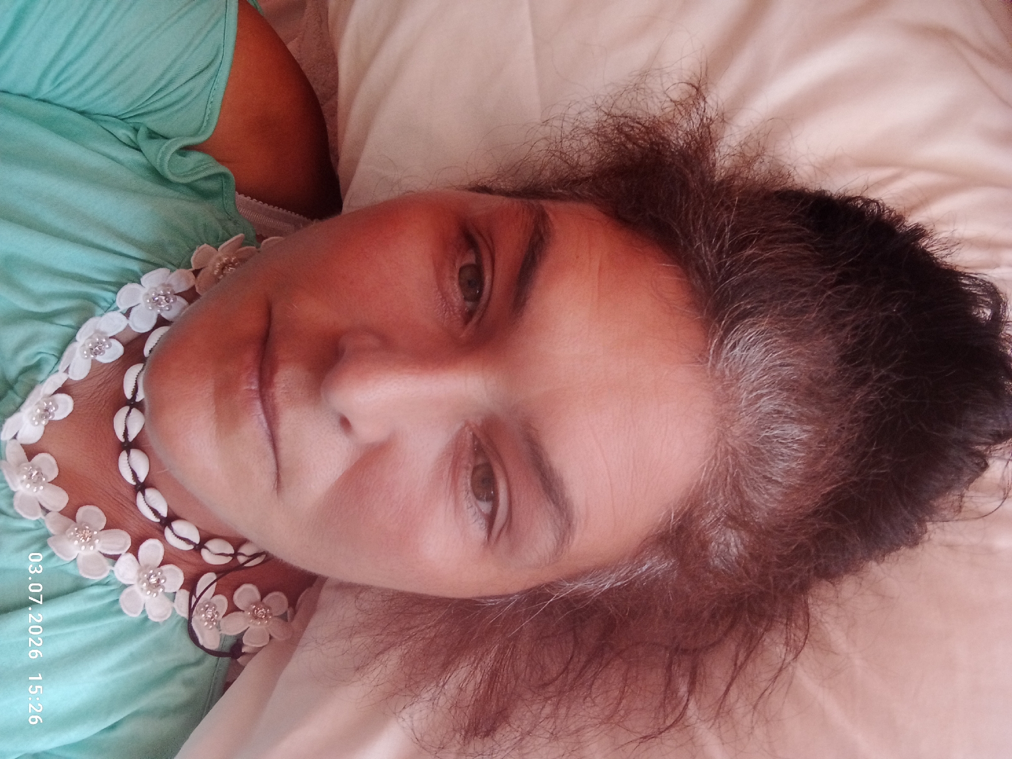This is a close-up, top-down, selfie-style portrait of a woman lying on a light-colored, wrinkled surface, possibly a bed or couch. The woman has medium-toned skin, with some gray hairs mixed in her dark, somewhat messy hair, which is pulled up. She is wearing a light teal, ruffled off-the-shoulder top and a decorative necklace made of white fabric flowers with small rhinestones and small cowrie shells. The lighting appears warm, possibly from natural light. A timestamp in the lower right corner reads "03.07.2026 15:26". There is no visible indication of the city of Ardea, Italy within the image itself.